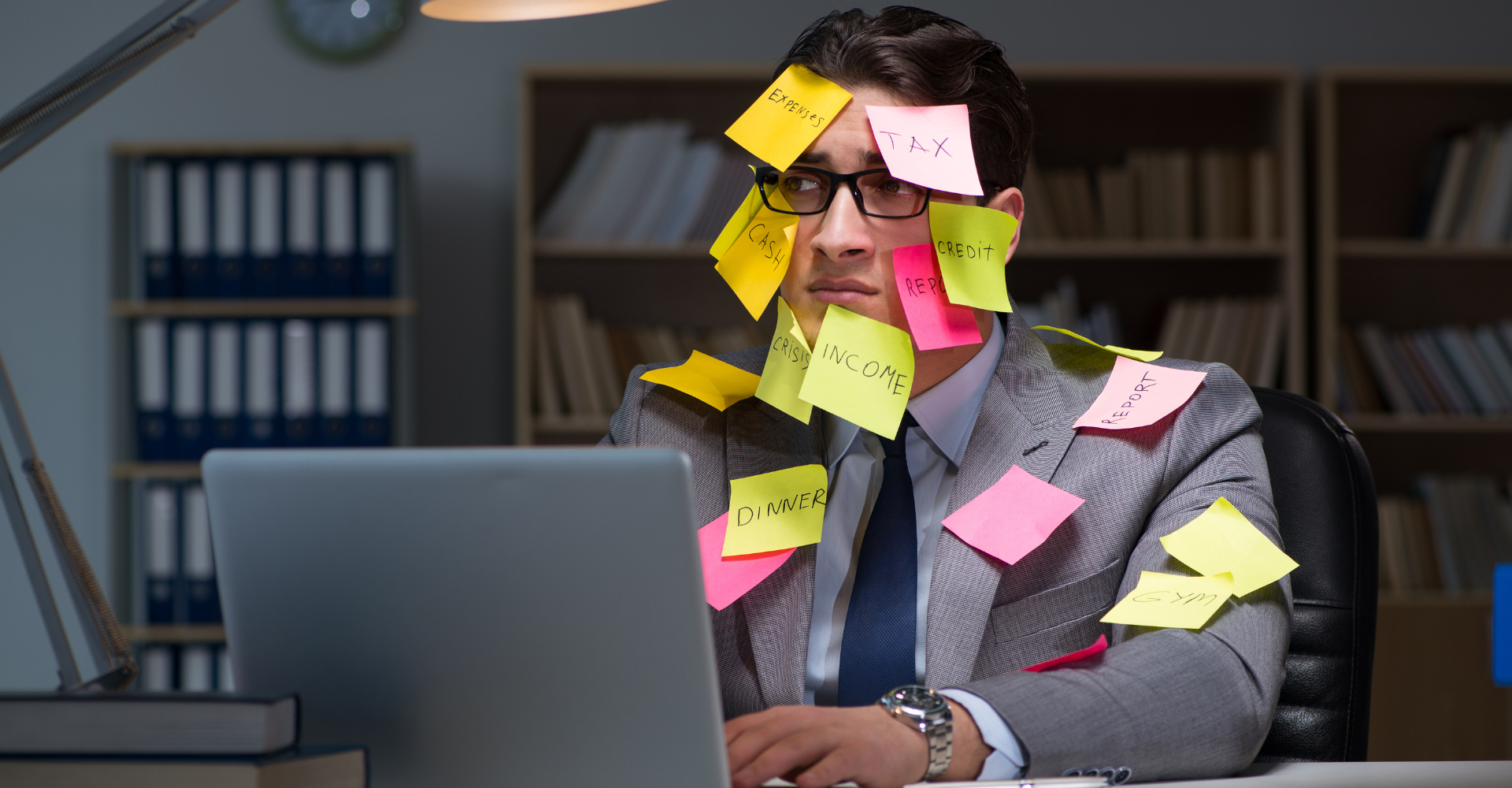 A man in a grey suit with a lot of colorful sticky notes on his face and upper body, typing at a computer screen.