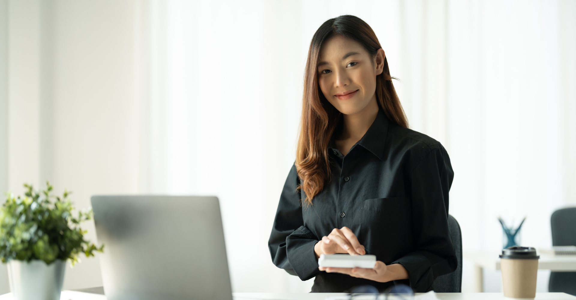 Smiling woman in a black shirt uses a smartphone at a white desk with a laptop, potted plant, and coffee cup.