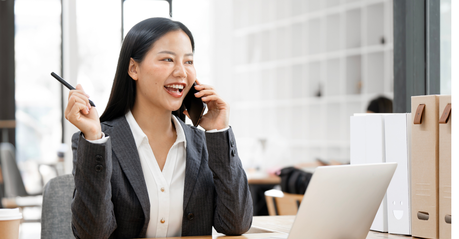 A woman in a business suit sits at a desk, smiling and talking on the phone. She holds a pen and has a laptop open.