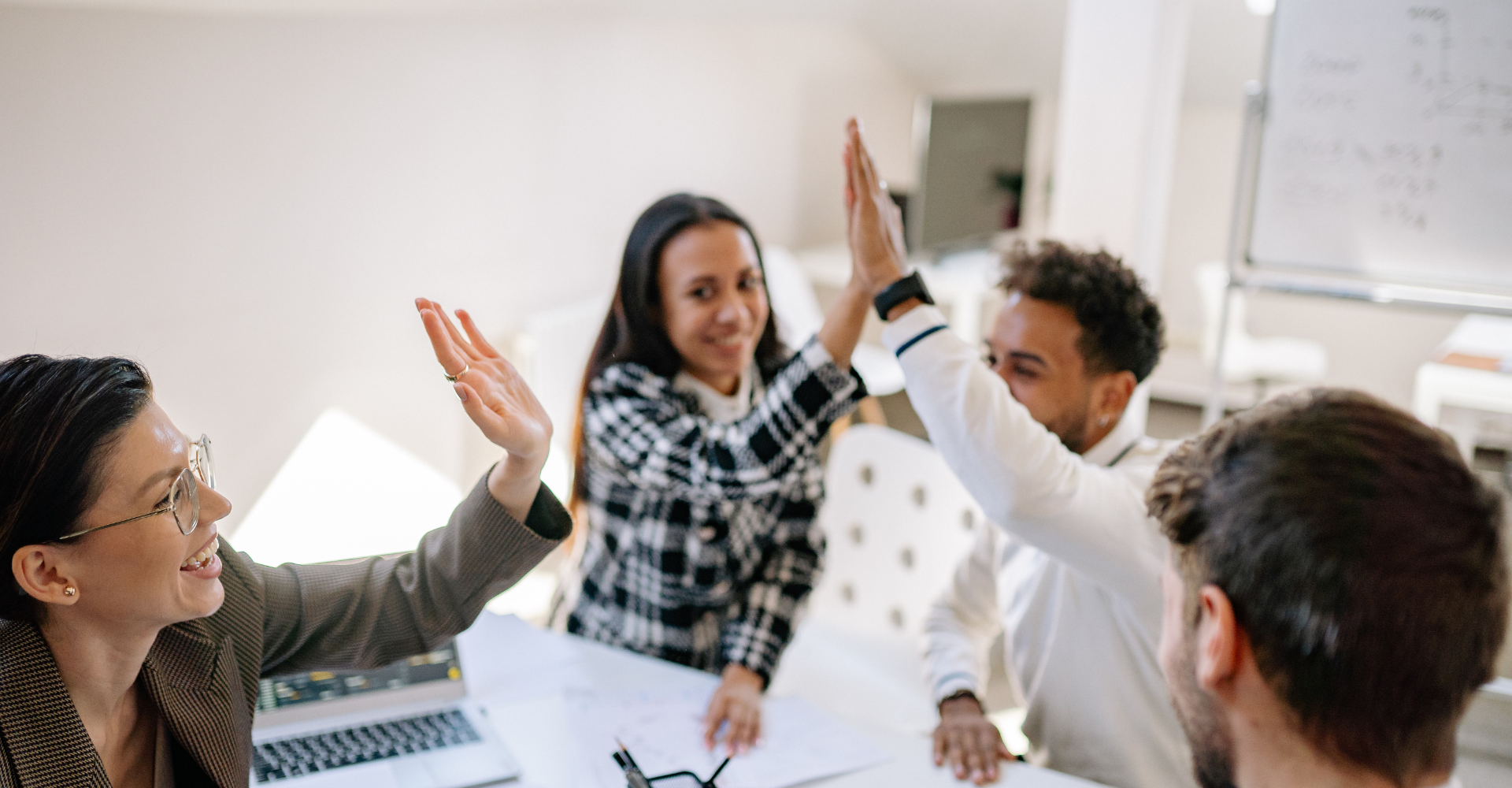 A group of four diverse colleagues at a meeting table, smiling and giving high-fives, creating an atmosphere of success.