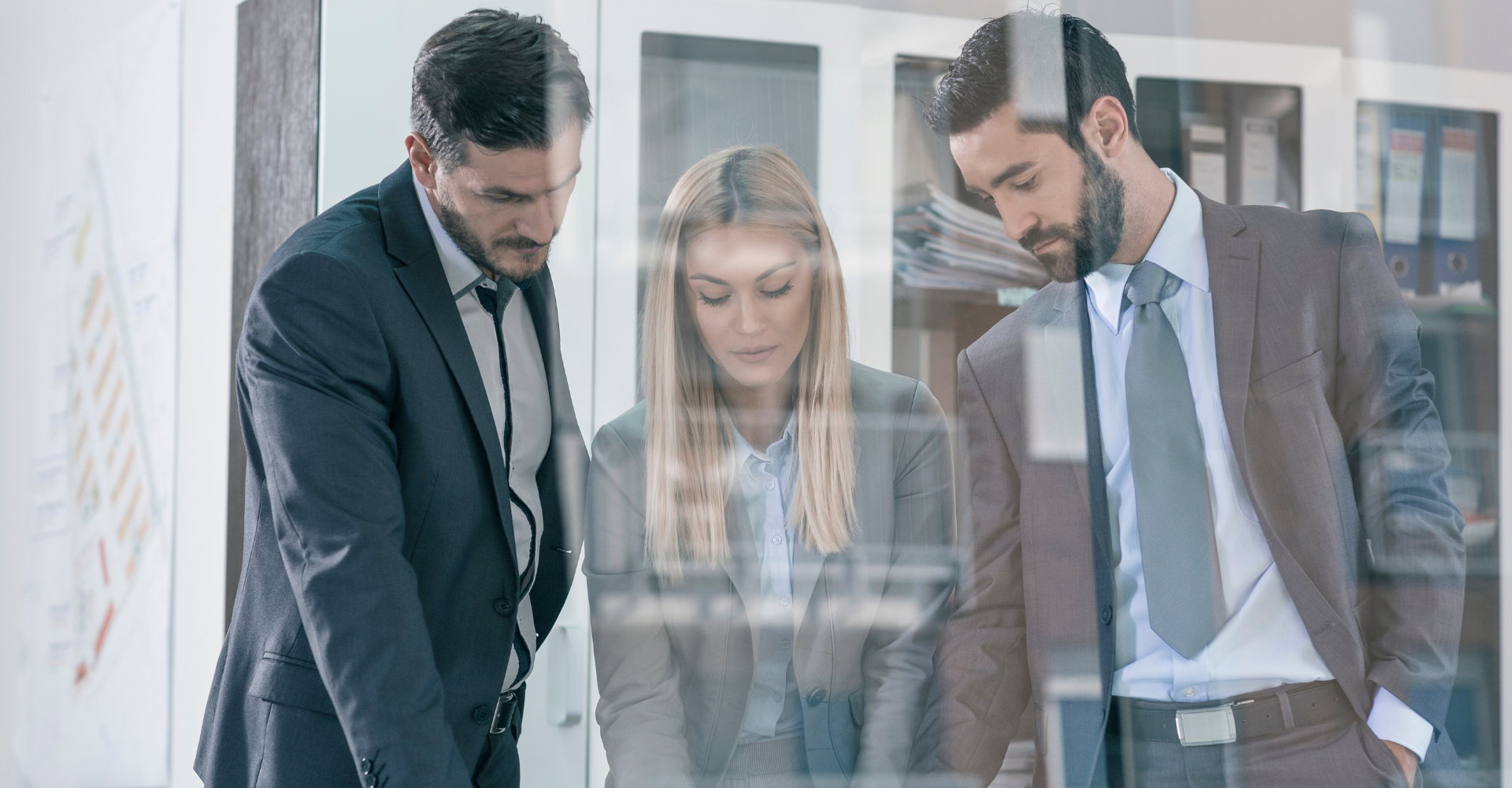 Three business professionals, two men and a woman, in a modern office, focused on documents. They appear serious.