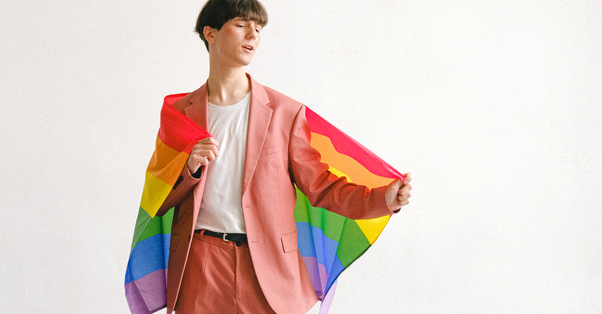 A person in a pink suit, holding a rainbow pride flag, appearing joyful and confident.