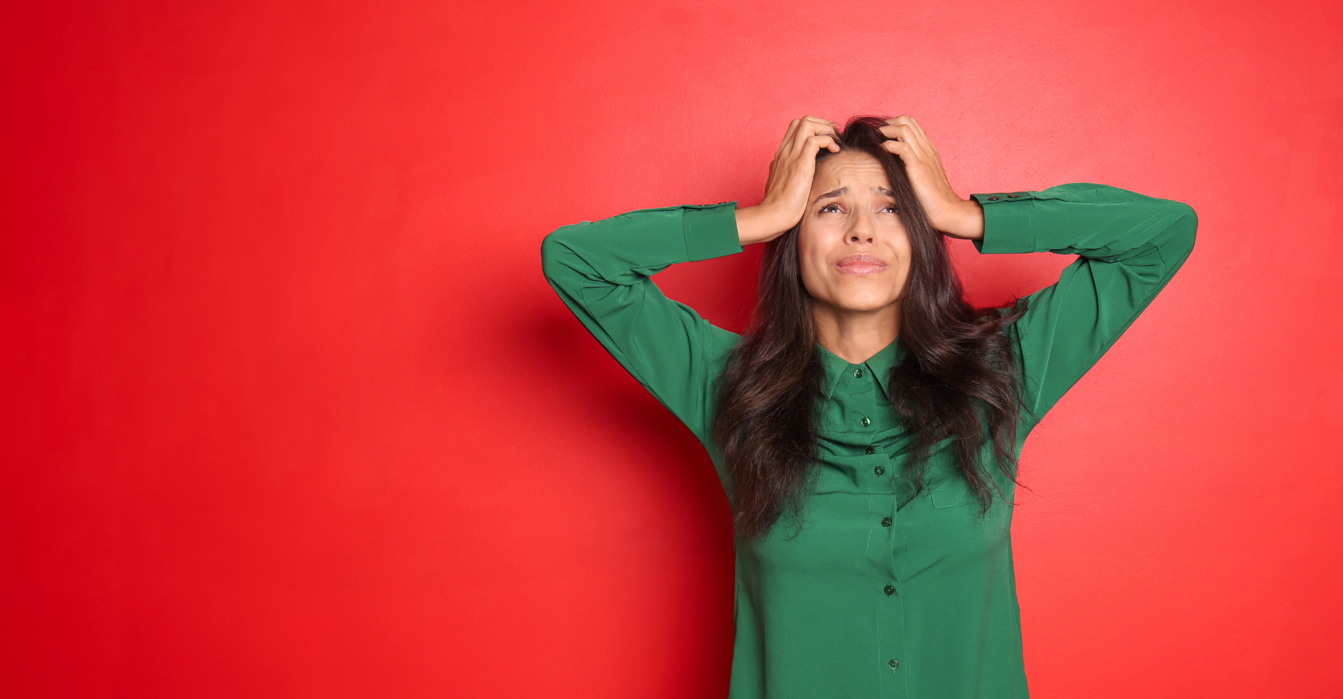 A woman in a green shirt stands against a red background, looking upwards with her hands on her head, expressing stress.