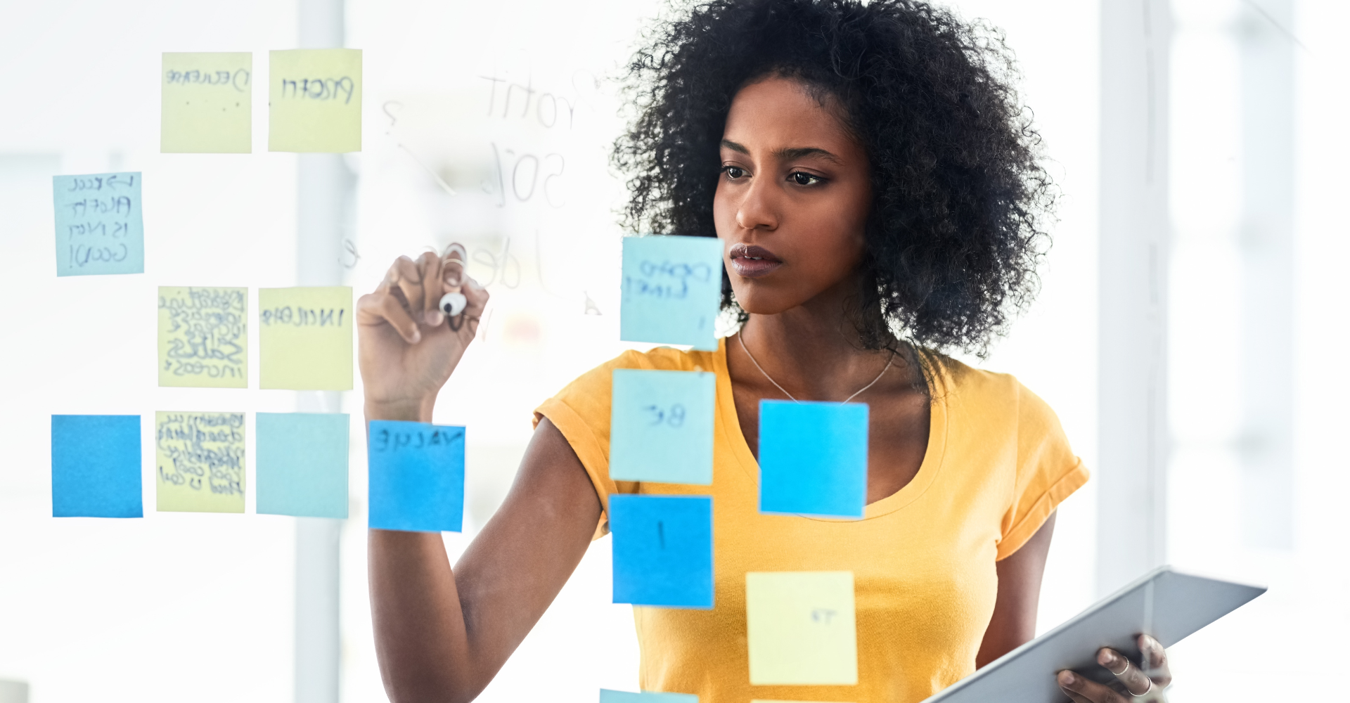 Woman in a yellow shirt writes on sticky notes arranged on a glass surface, holding a tablet. The scene conveys focus.