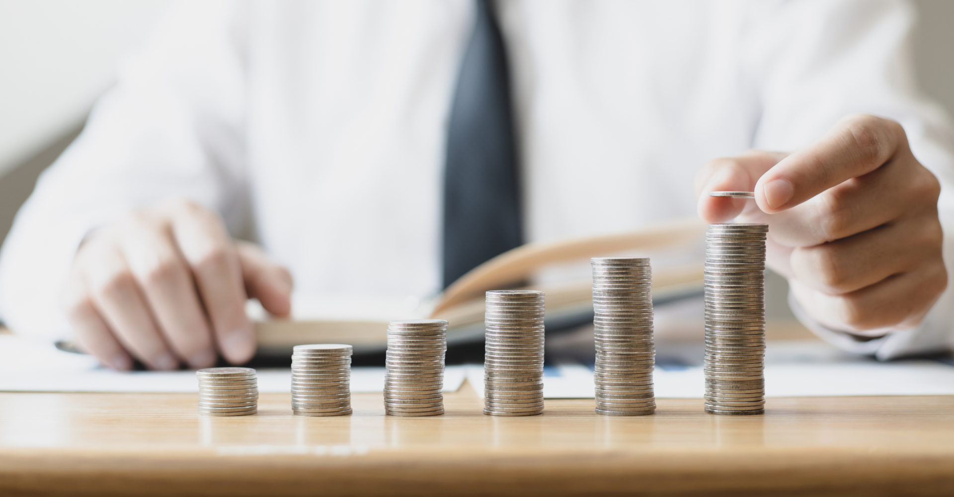 A person in a suit places a coin on one of six ascending stacks of coins on a desk.