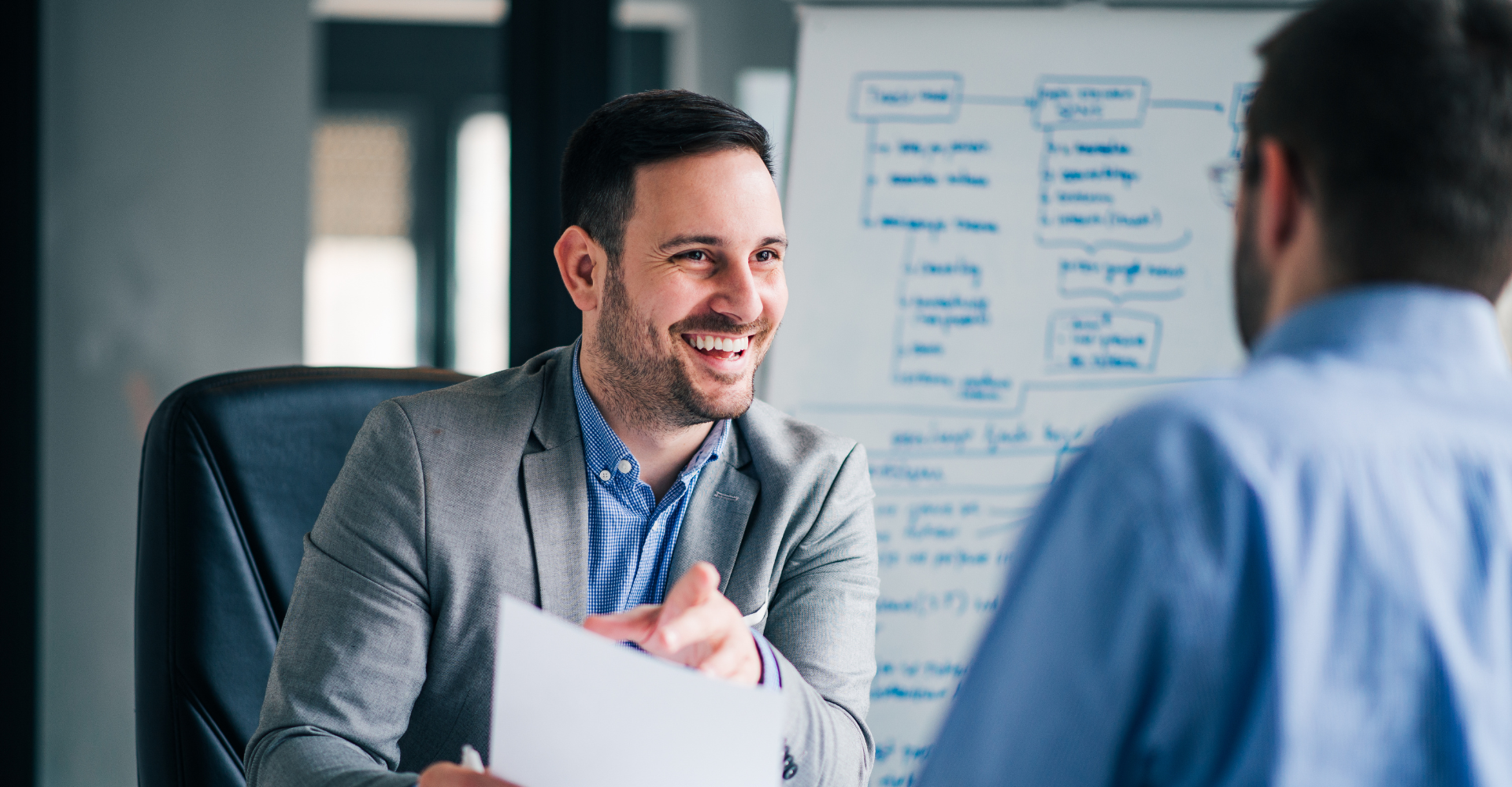 A man smiles and holds a paper during a discussion with a colleague. A whiteboard with flow charts is in the background.