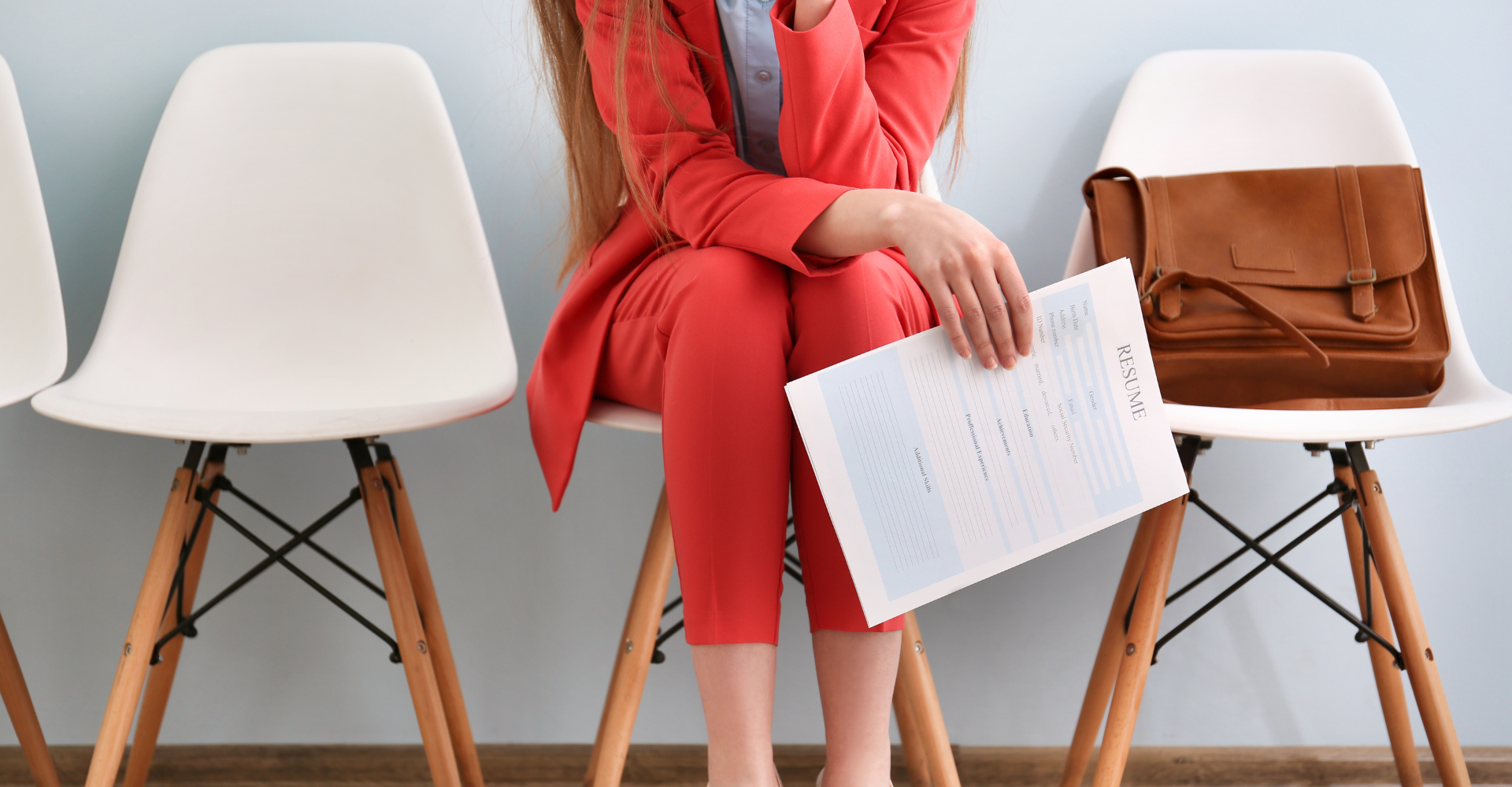 A person in a red suit sits anxiously on a white chair holding a resume, with a brown bag on the chair beside them.