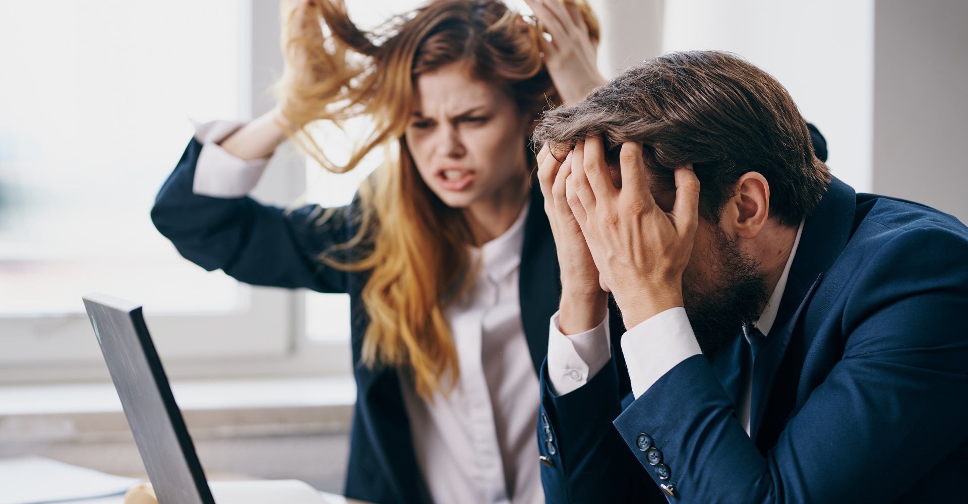 A frustrated woman pulls her hair beside a man with his head in his hands, both seated at a desk with a laptop.