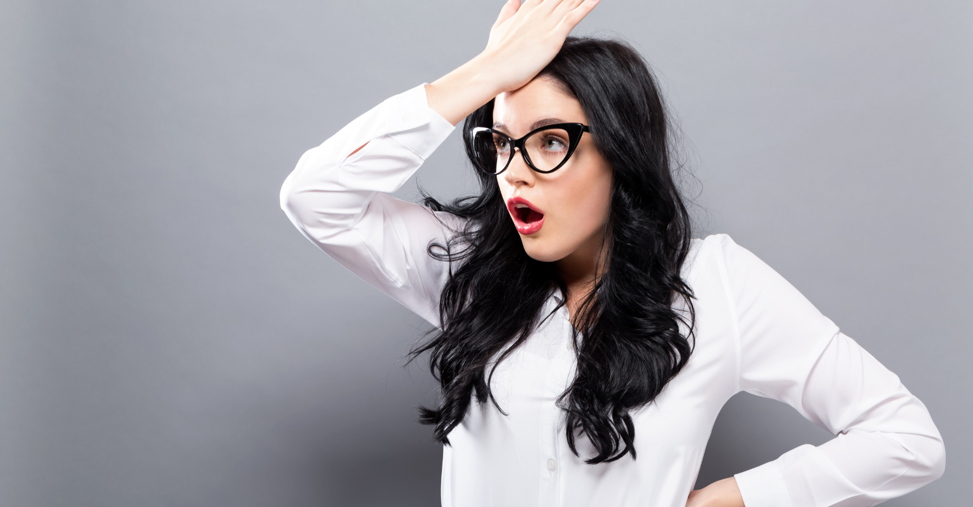 Surprised woman with long black hair and glasses, hand on forehead, against gray background, showing realization.