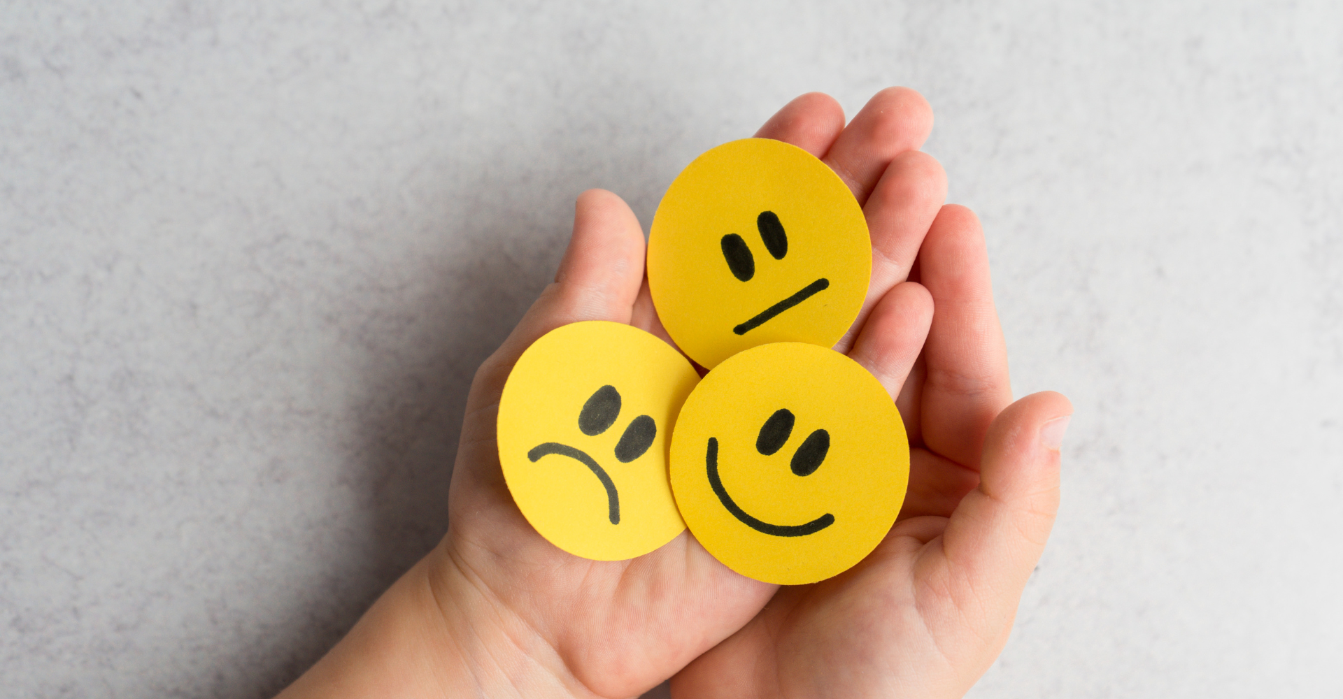Hands holding three yellow paper circles with black drawn faces: one smiling, one neutral, and one sad.