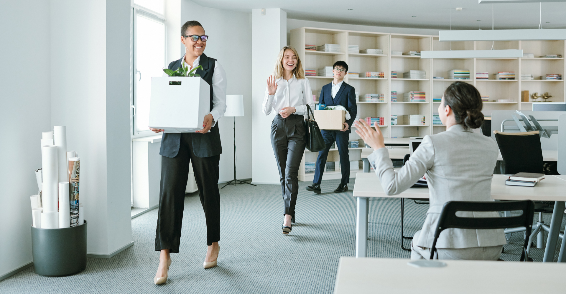 Three people carrying boxes and smiling enter an office. Colleagues greet them with waves, creating an atmosphere of warmth.