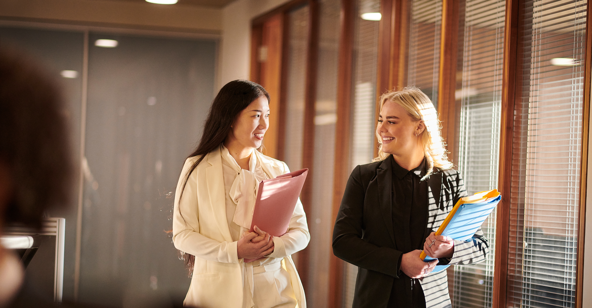 Two businesswomen walk in an office corridor, chatting and smiling. One holds a pink folder, the other orange and blue.