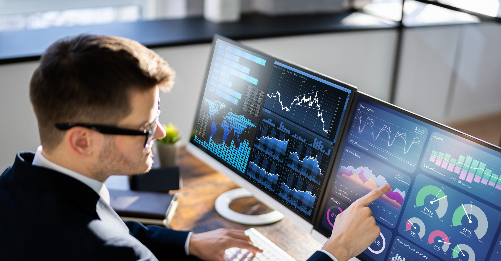 A man with glasses sitting at a desk, pointing at a screen where there are many different graphs.