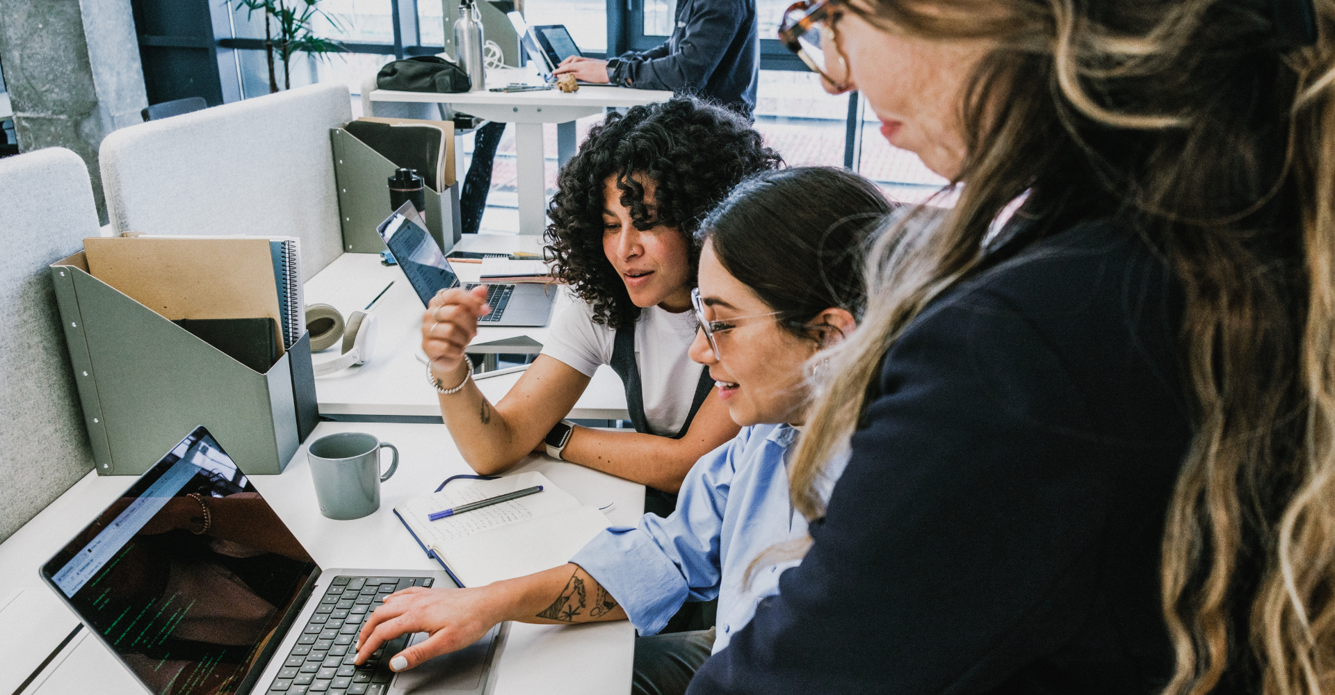 Three people collaborate at a modern office desk, focusing on a laptop screen. The mood is lively and focused.
