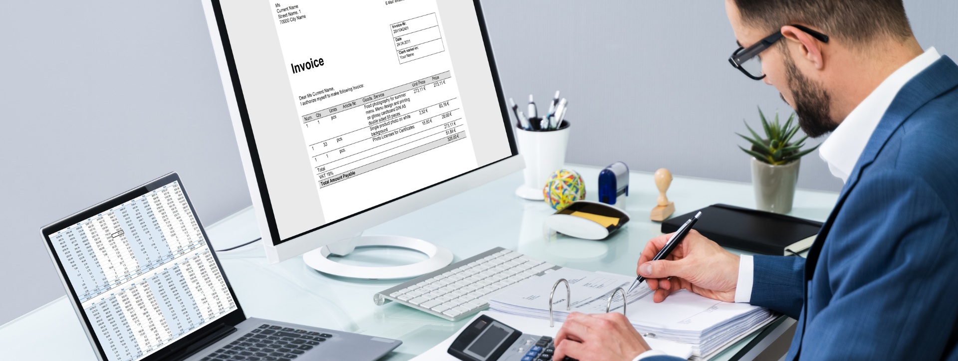 Man in an office analyzes documents at a desk with a large monitor displaying an invoice and a laptop showing a spreadsheet.