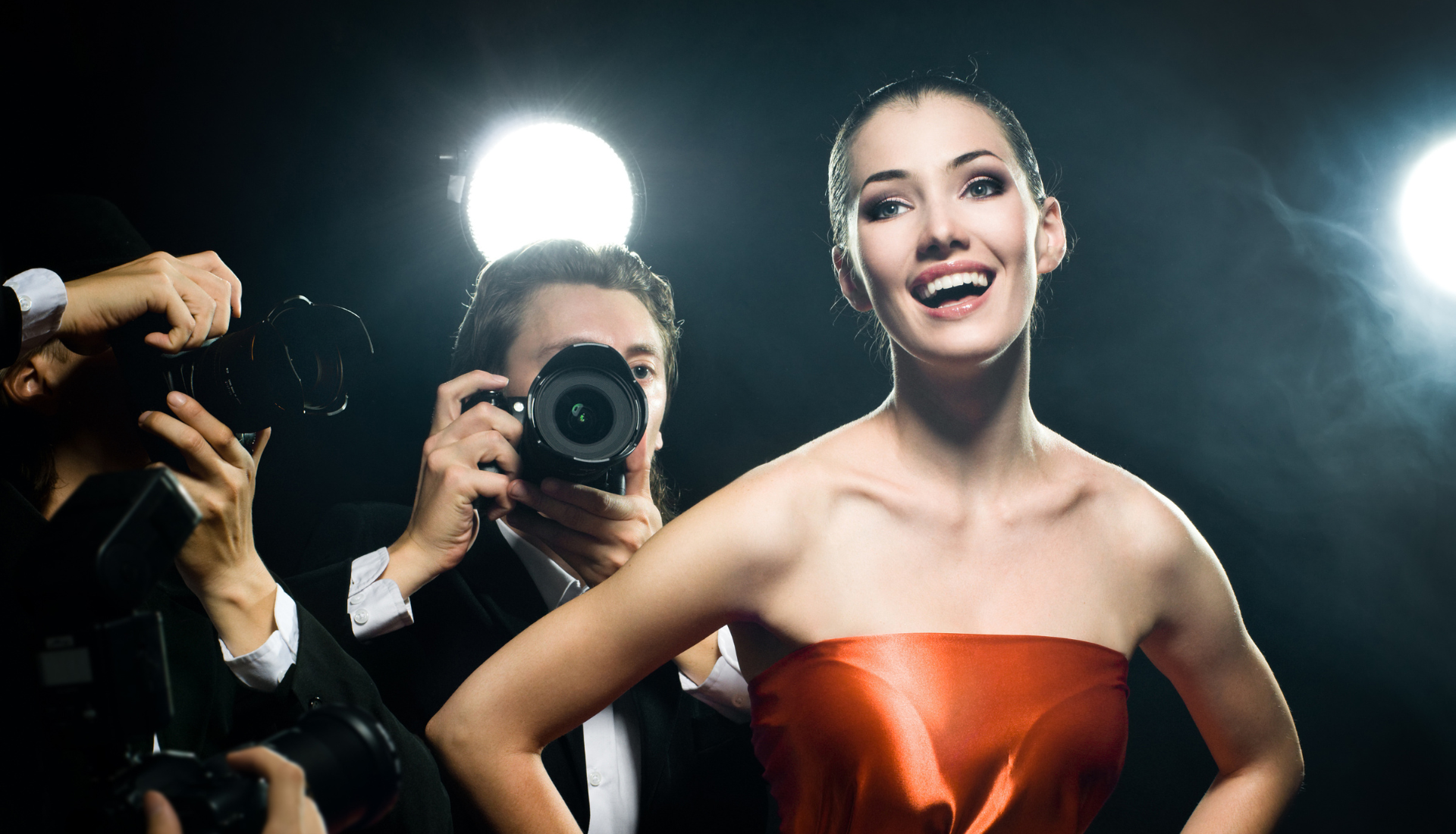 A woman wearing a strapless red dress in a black room with studio lights, being photographed by 2 men in suits behind her