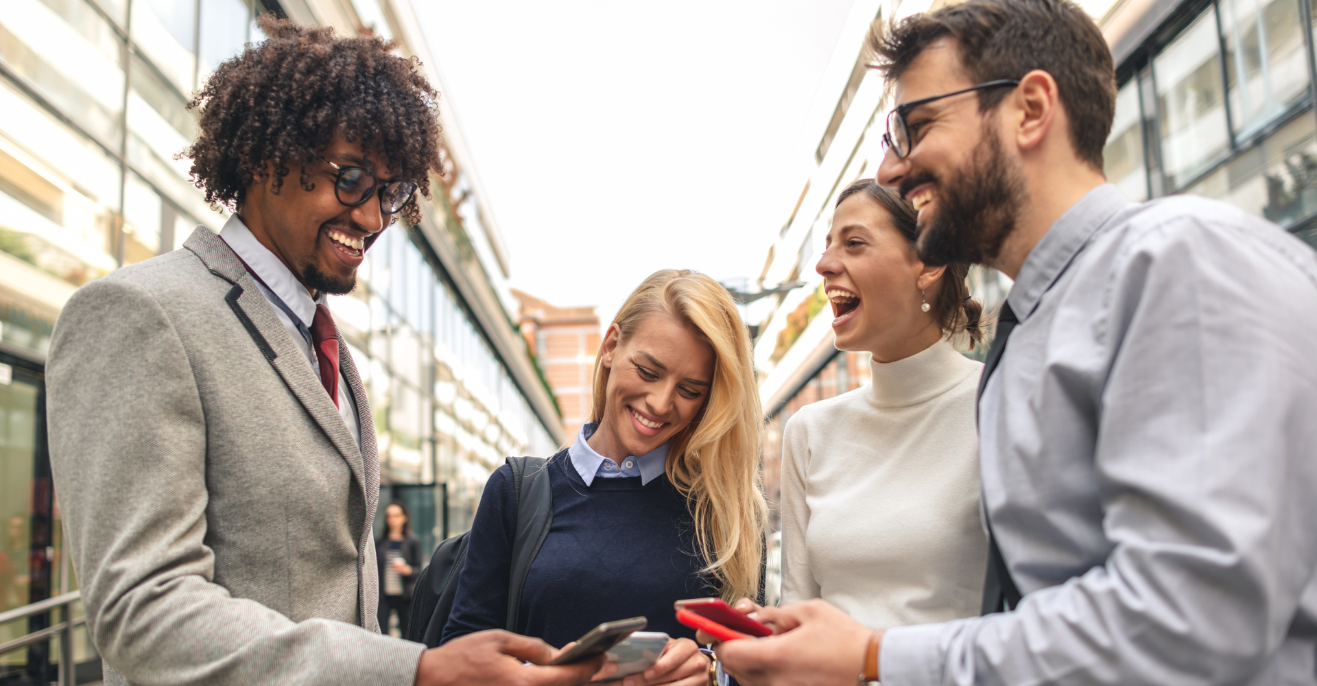 A diverse group of four people stand outdoors, smiling and interacting while looking at smartphones.
