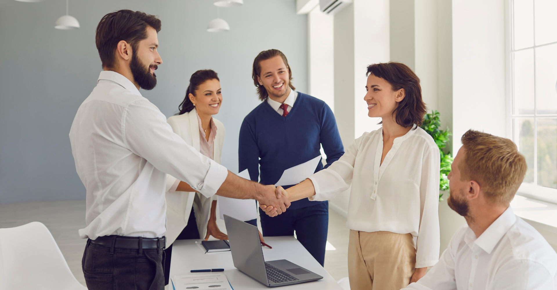 People in an office, two shaking hands with smiles, others observing with approval. Laptops and papers are on the table.