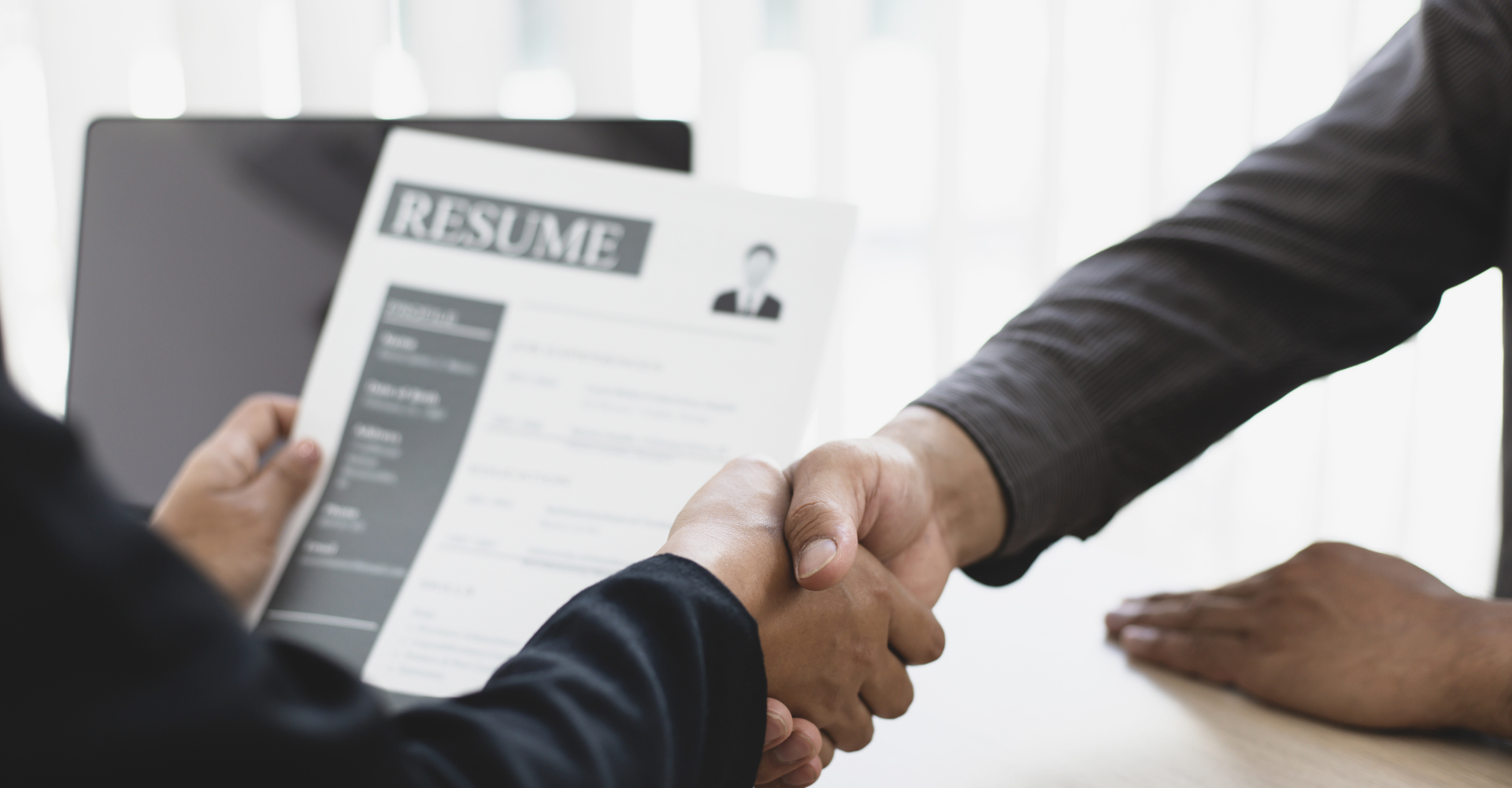 Two people shaking hands over a table, one holding a resume. The scene conveys a successful job interview.