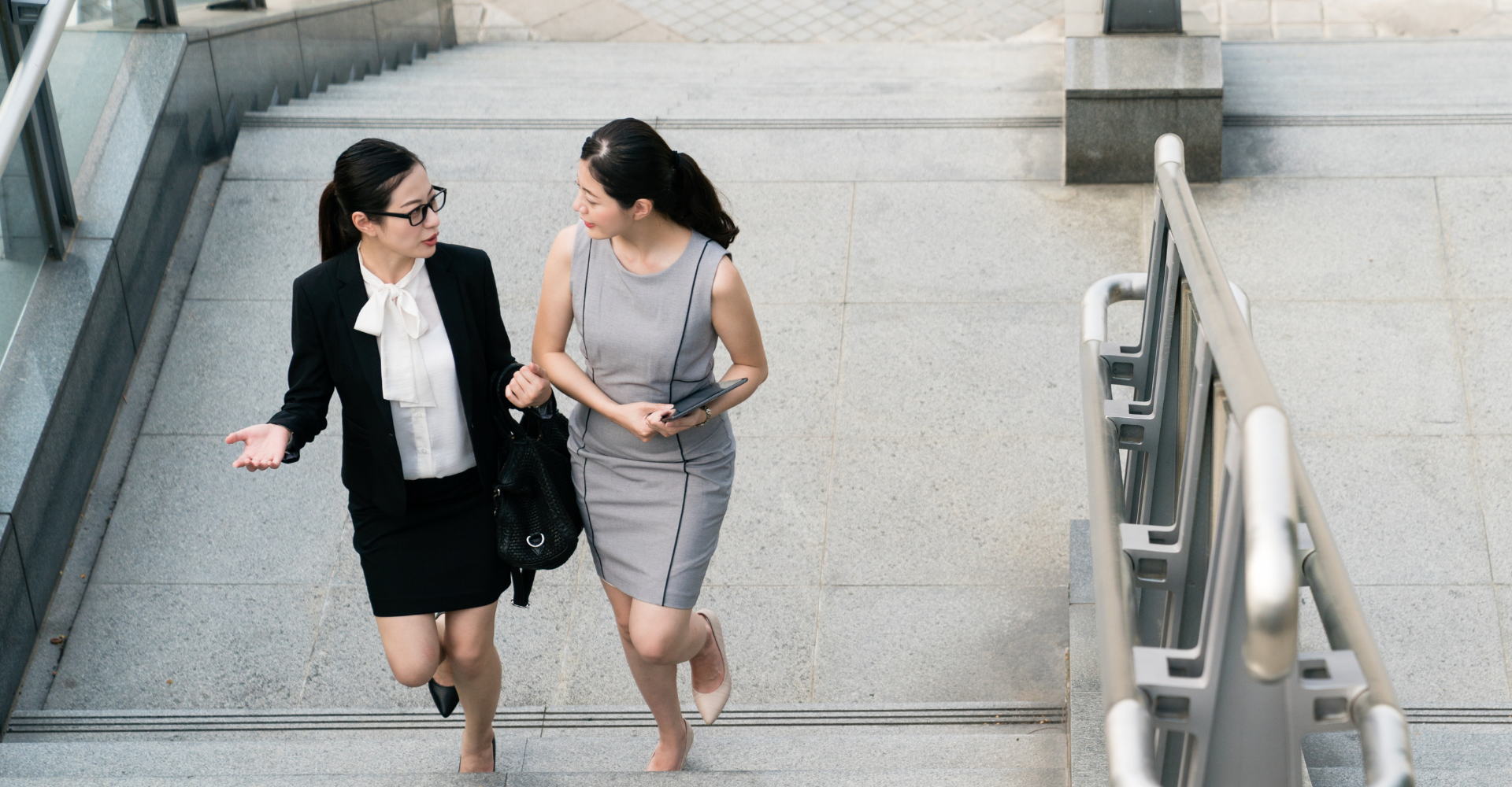 Two professional women walk up stairs, engaged in conversation. One wears a black suit, the other a gray dress.