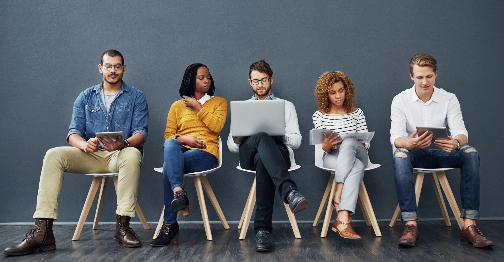 A diverse group of five people sits on chairs, holding digital devices. They appear focused and engaged in their screens.