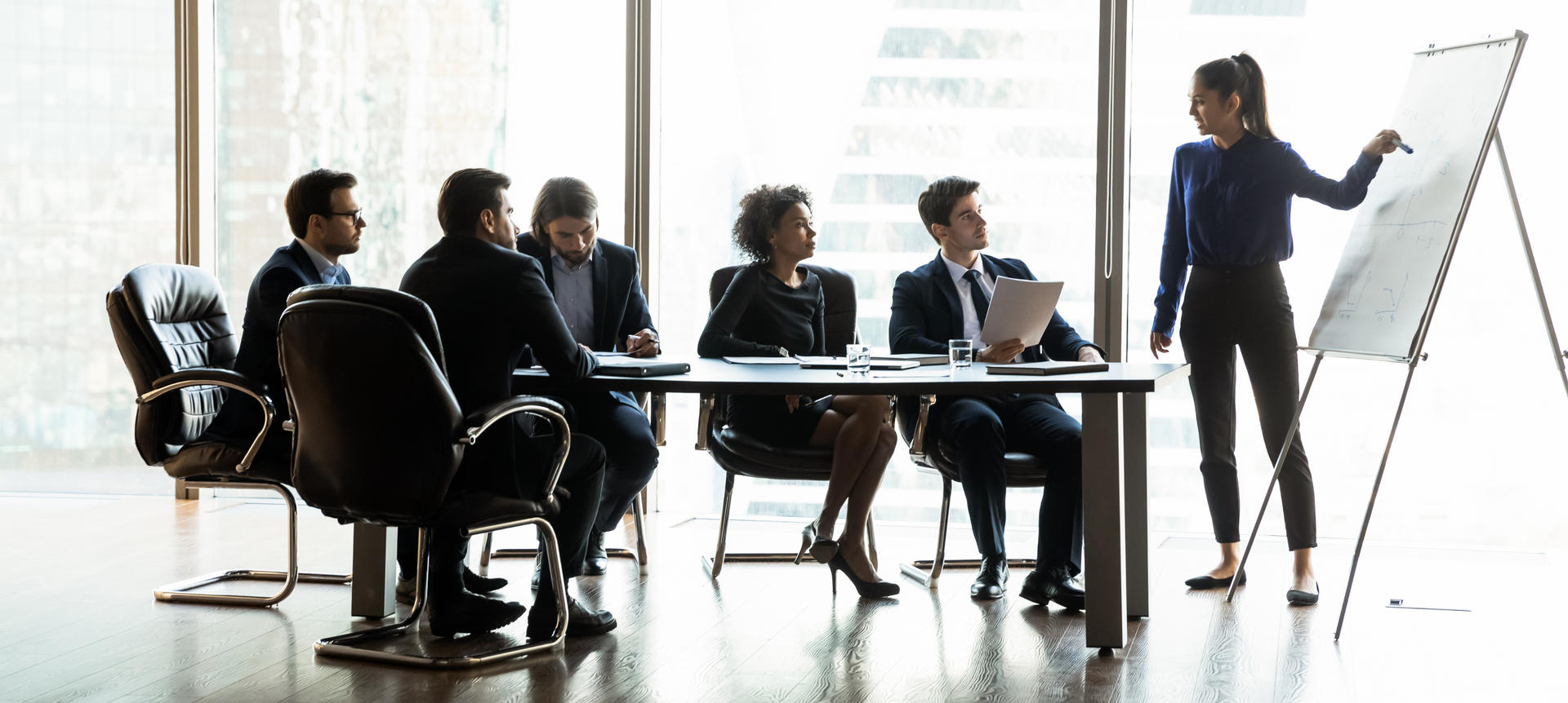 Five business people sitting around a table, looking towards a woman standing, giving a presentation.