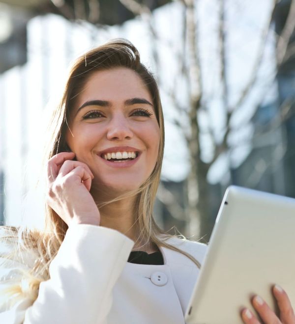 Smiling woman wearing a white coat outdoors holding a tablet in one hand and a phone to her ear.