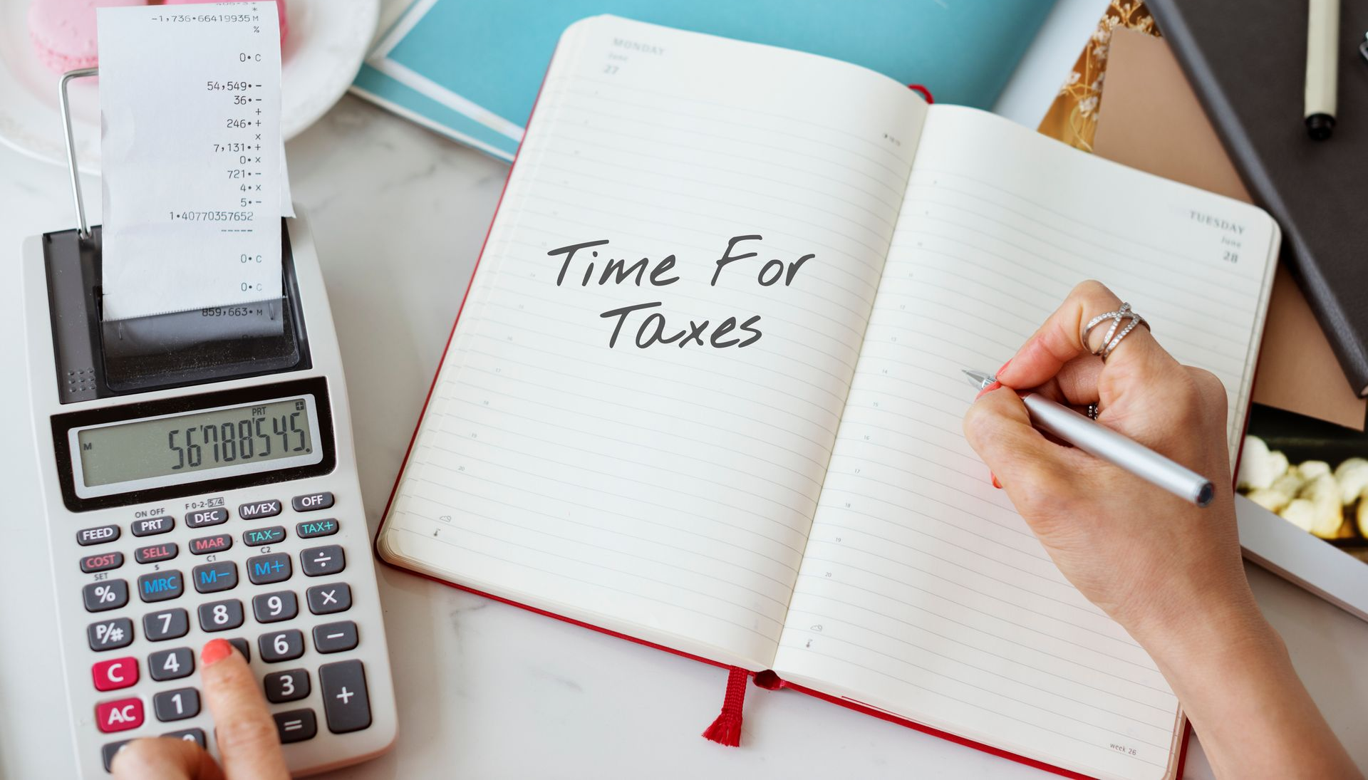A hand writes "Time For Taxes" in an open planner. Nearby, another hand uses a calculator with a printed receipt.
