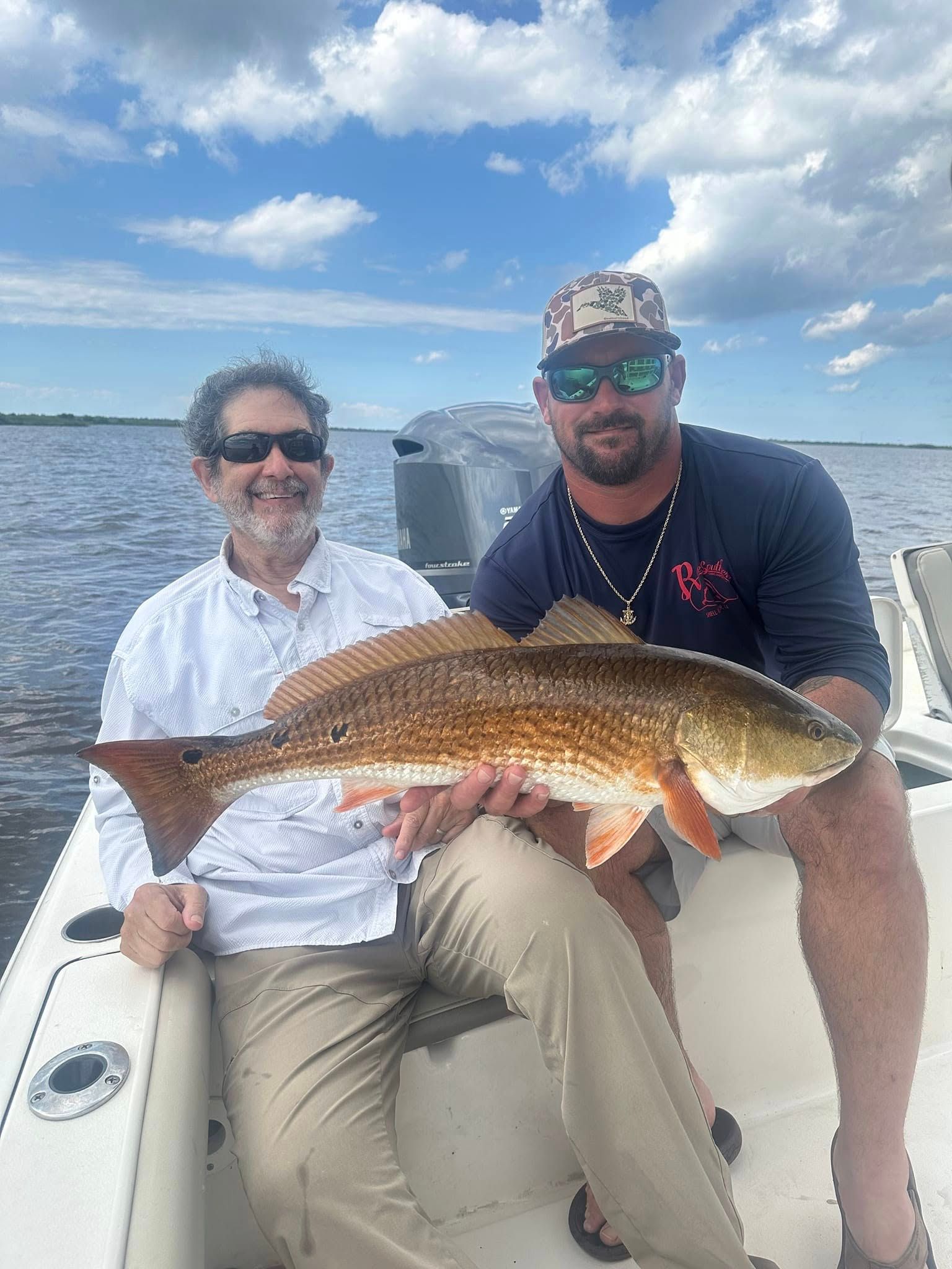Two people smiling on a boat, holding a large redfish caught in open water under a cloudy blue sky.