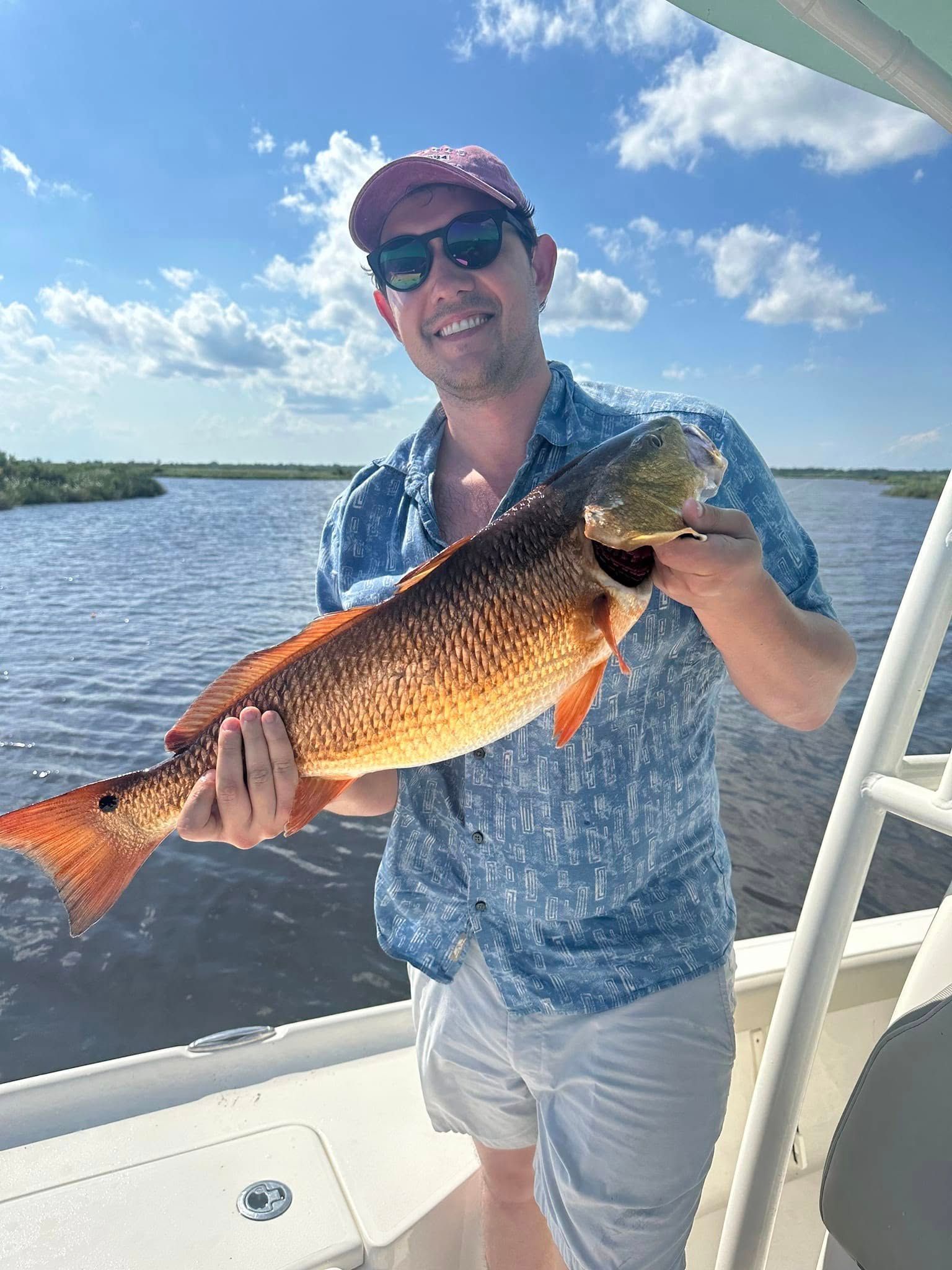 A person wearing a patterned blue shirt, shorts, and a cap holds a large, coppery-colored redfish on a boat near marshland.