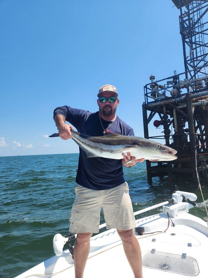A person wearing a hat and sunglasses holding a large fish on a boat next to an offshore platform.