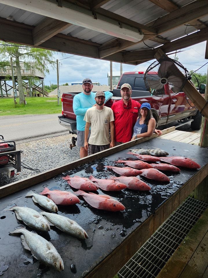 Four people standing behind a fish-cleaning table filled with freshly caught red snapper and smaller fish, near a truck.