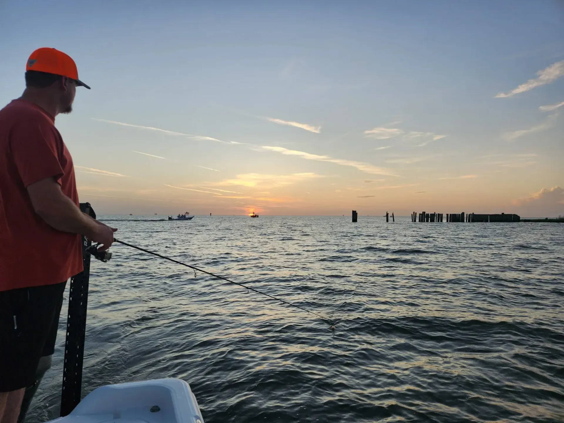 A person in a bright orange hat and shirt stands on a boat at sunset, holding a fishing rod over the water.