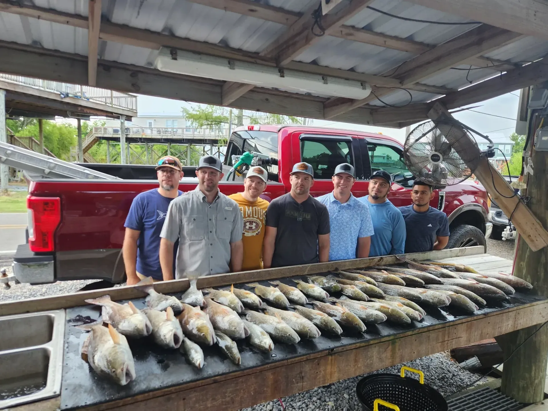 A group of eight people stand behind a long wooden table covered with a large catch of fish, with a red truck in the back.