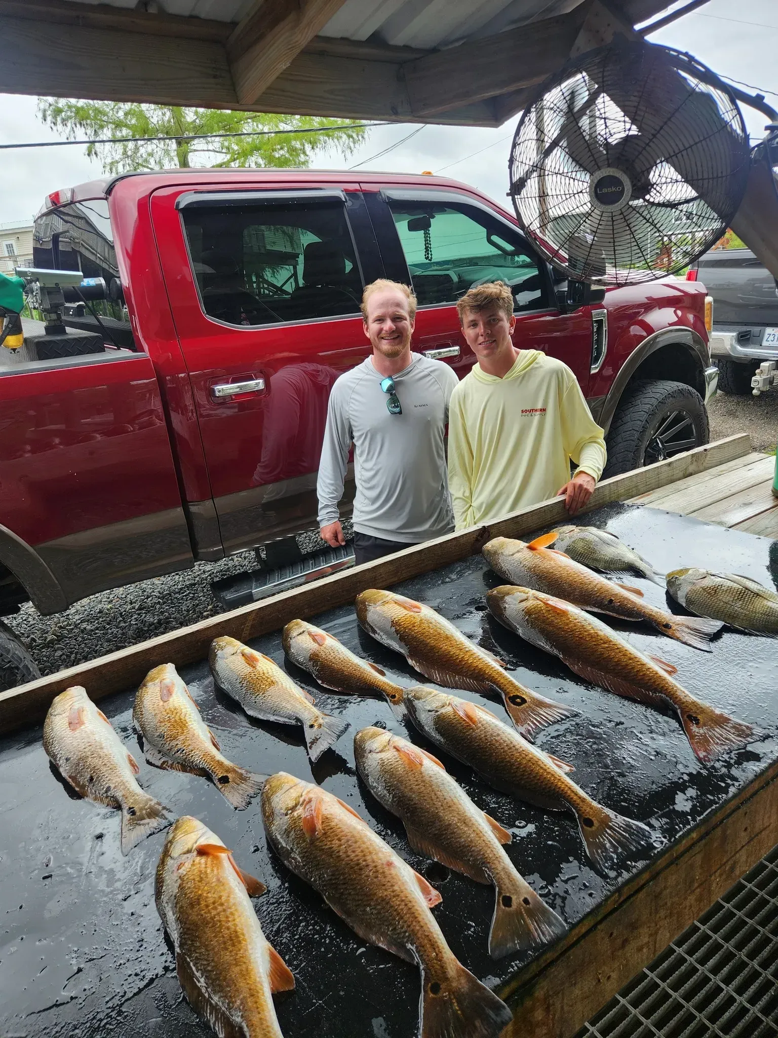 Two people stand smiling behind a table covered with freshly caught redfish, with a large red truck in the background.