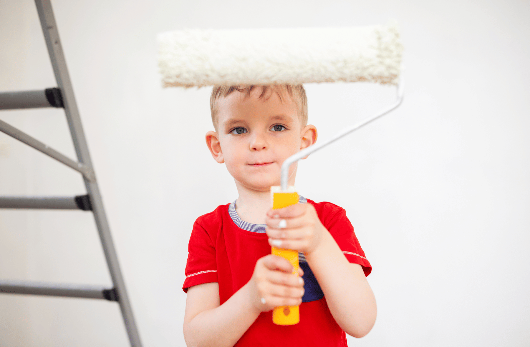 Child in a red shirt holding a paint roller near a white wall, with a ladder in the background