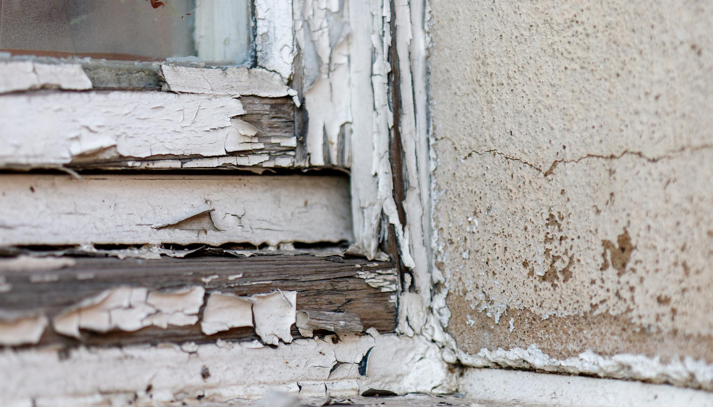 Close-up of peeling white paint on a wooden window frame and adjacent concrete wall.