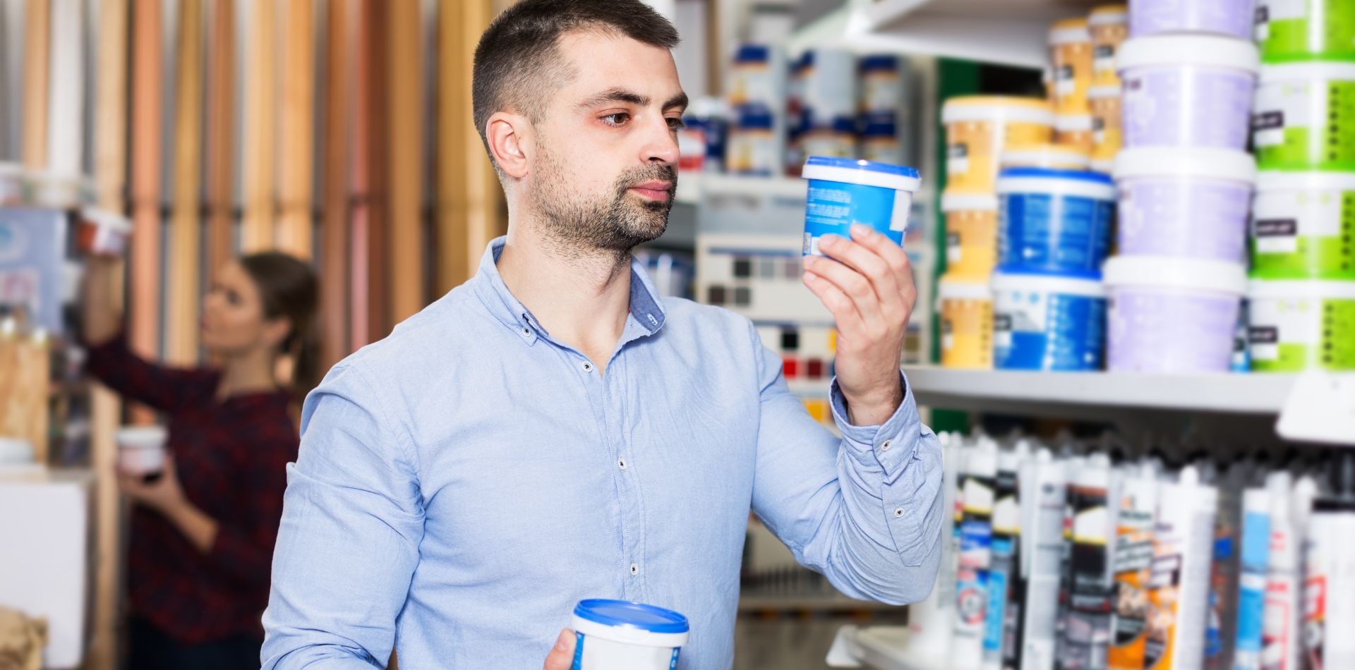 Man examining paint cans in a store, other person browsing in the background.