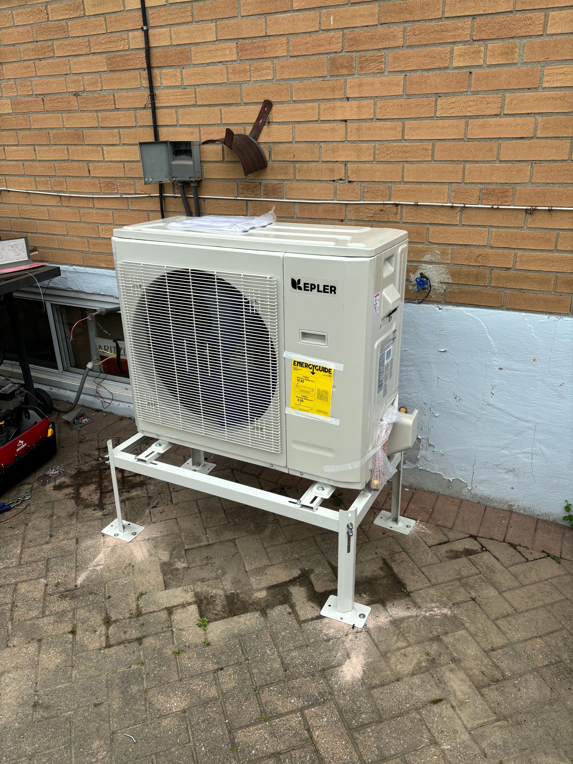 Air conditioning unit, mounted on a white stand against a brick wall.