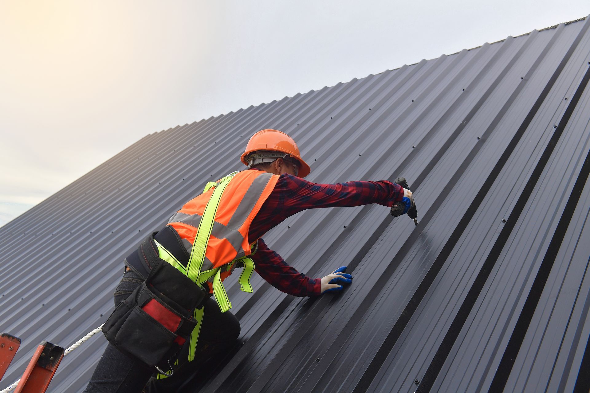 Worker in orange safety gear climbing a steep corrugated metal roof with a harness