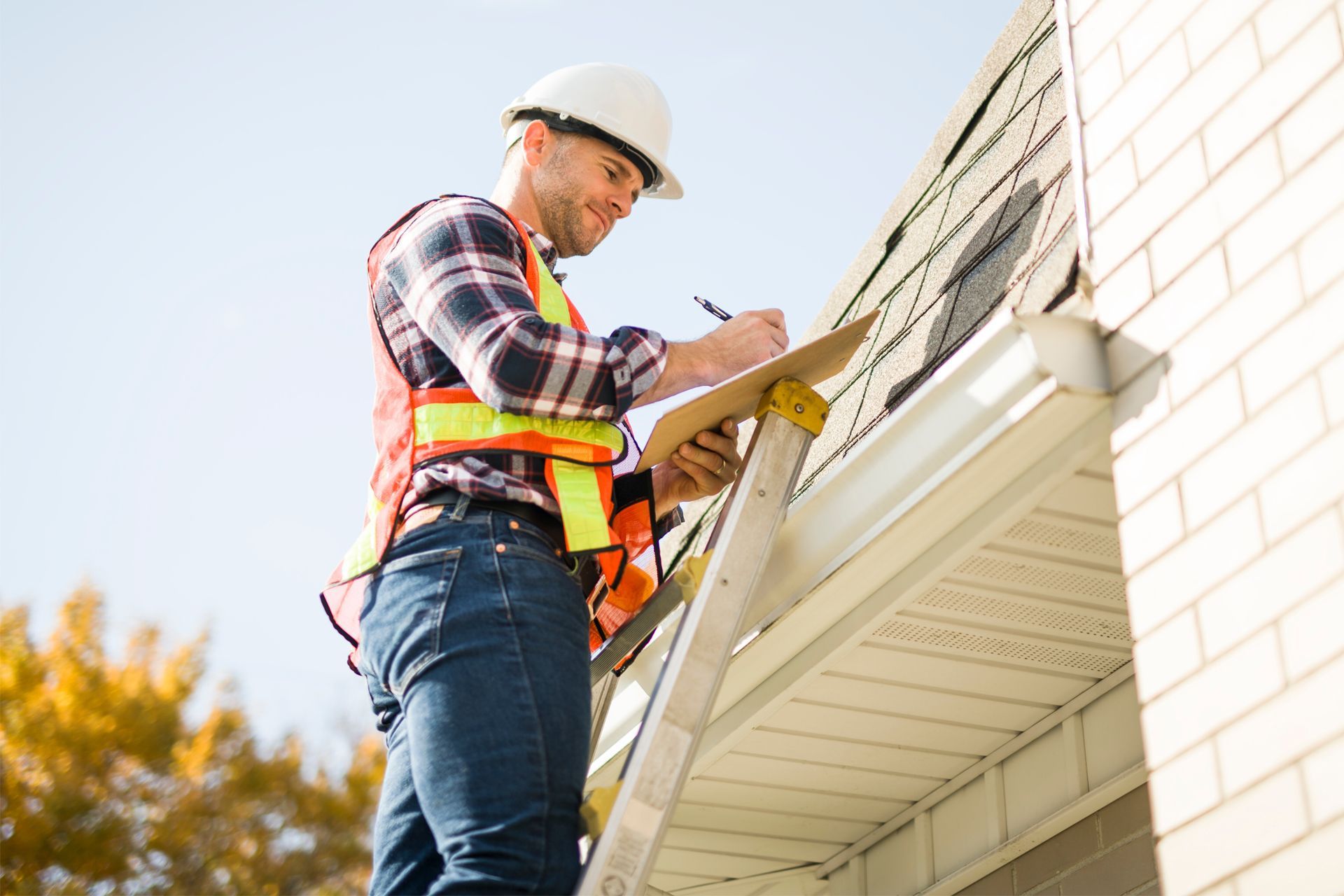 Worker in hard hat and safety vest checks notes on a ladder beside a house roofline.