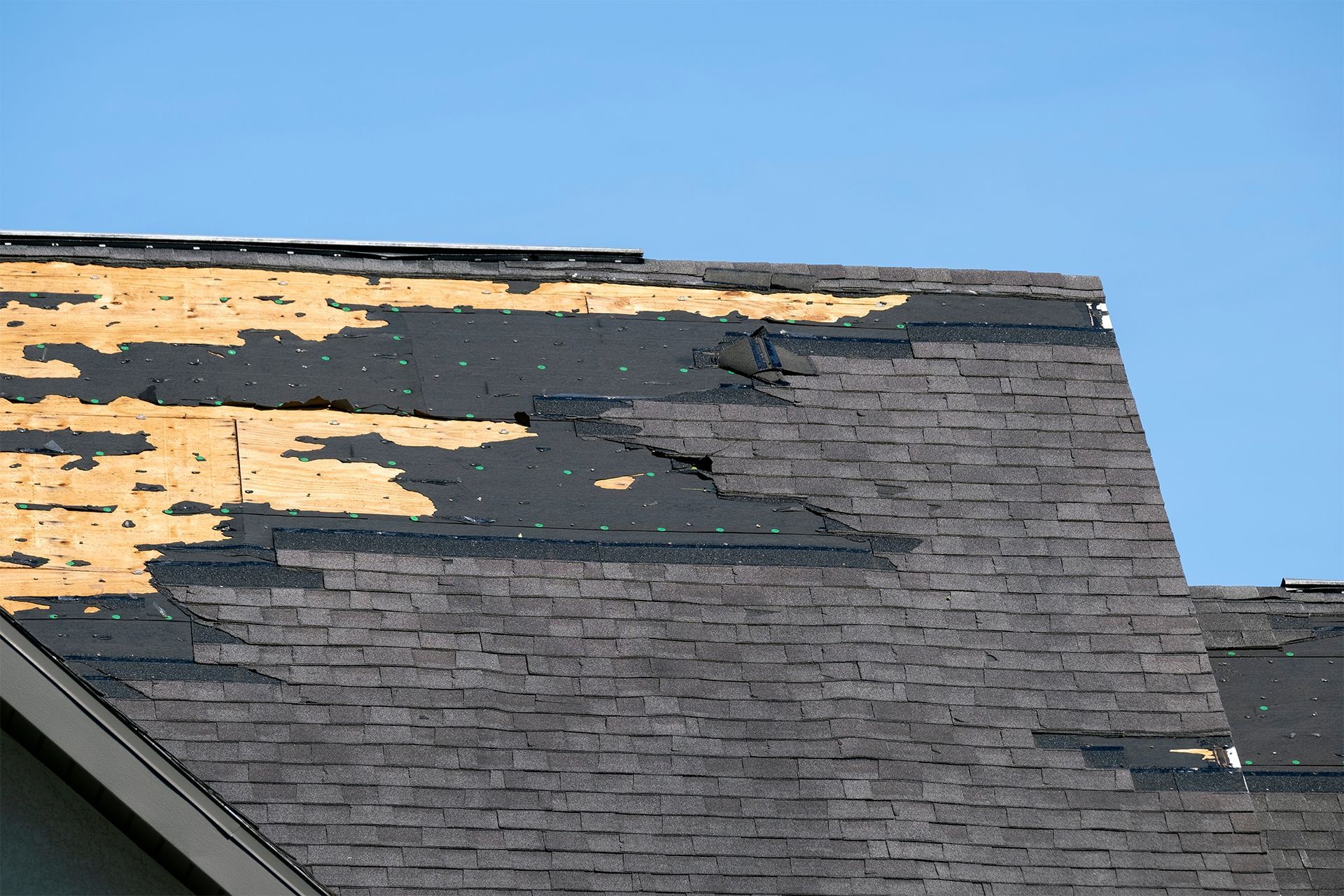 Damaged shingled roof with missing black and tan shingles against a blue sky.