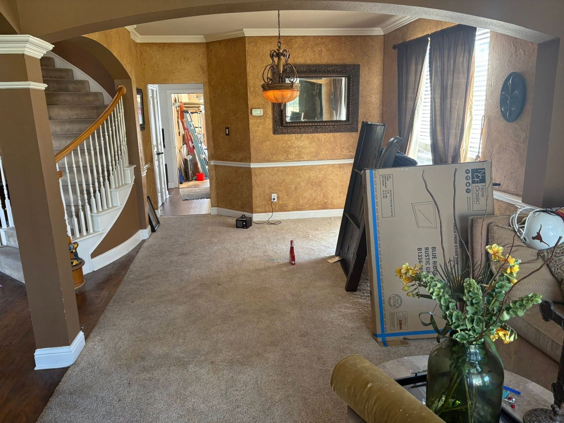 Carpeted living room hallway with stairs, window blinds, and flowers on a table; a large panel leans by the wall.