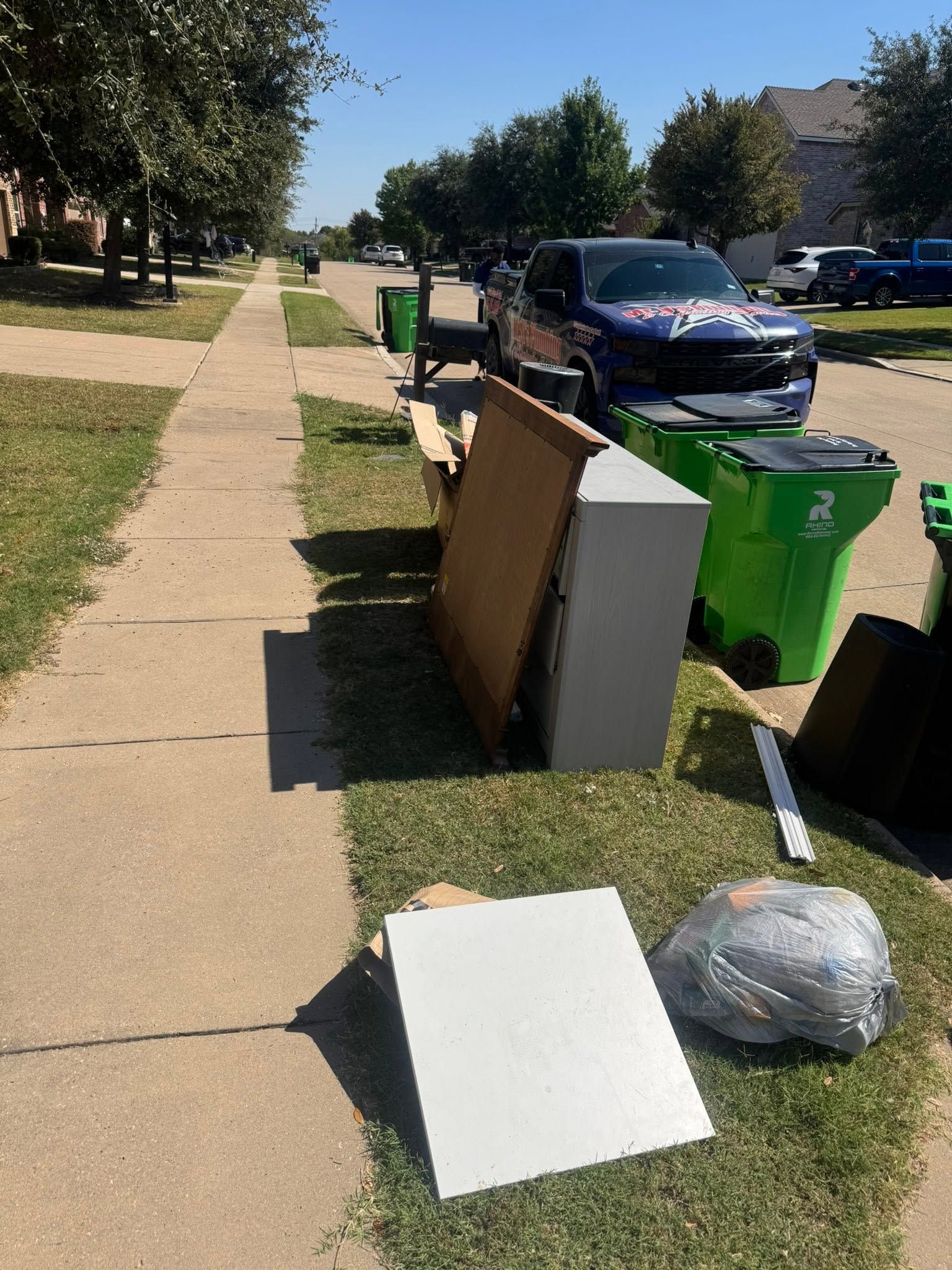 Sidewalk lined with curbside trash, cardboard, and green bins beside parked cars on a sunny street