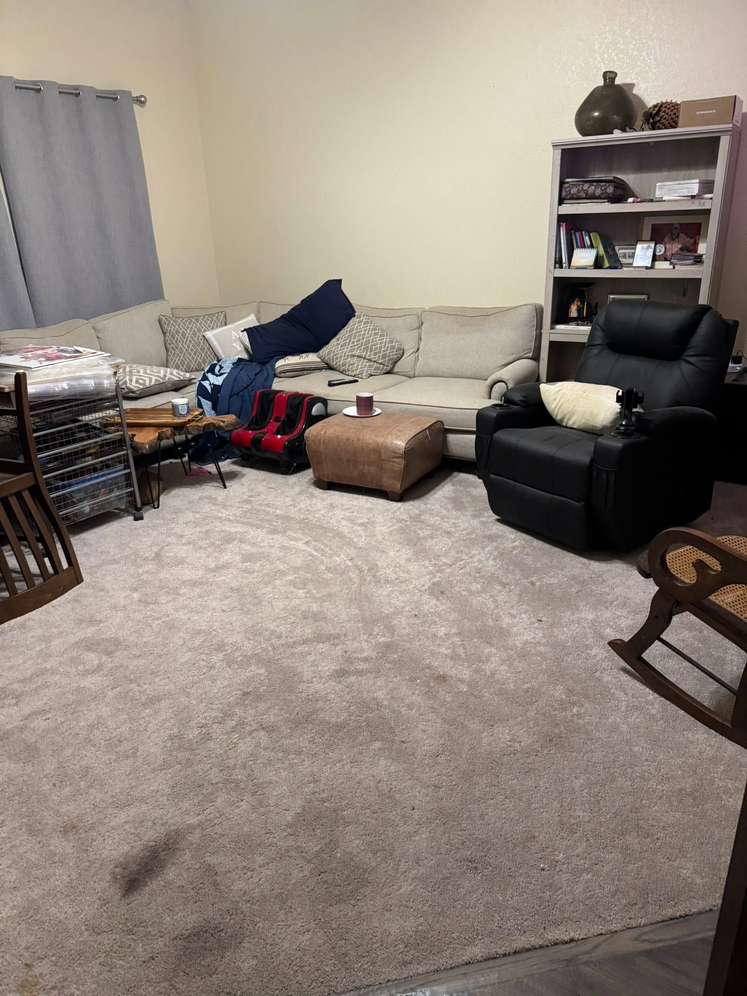 Cluttered living room with beige carpet, gray couch, black recliner, shelves, and scattered items.