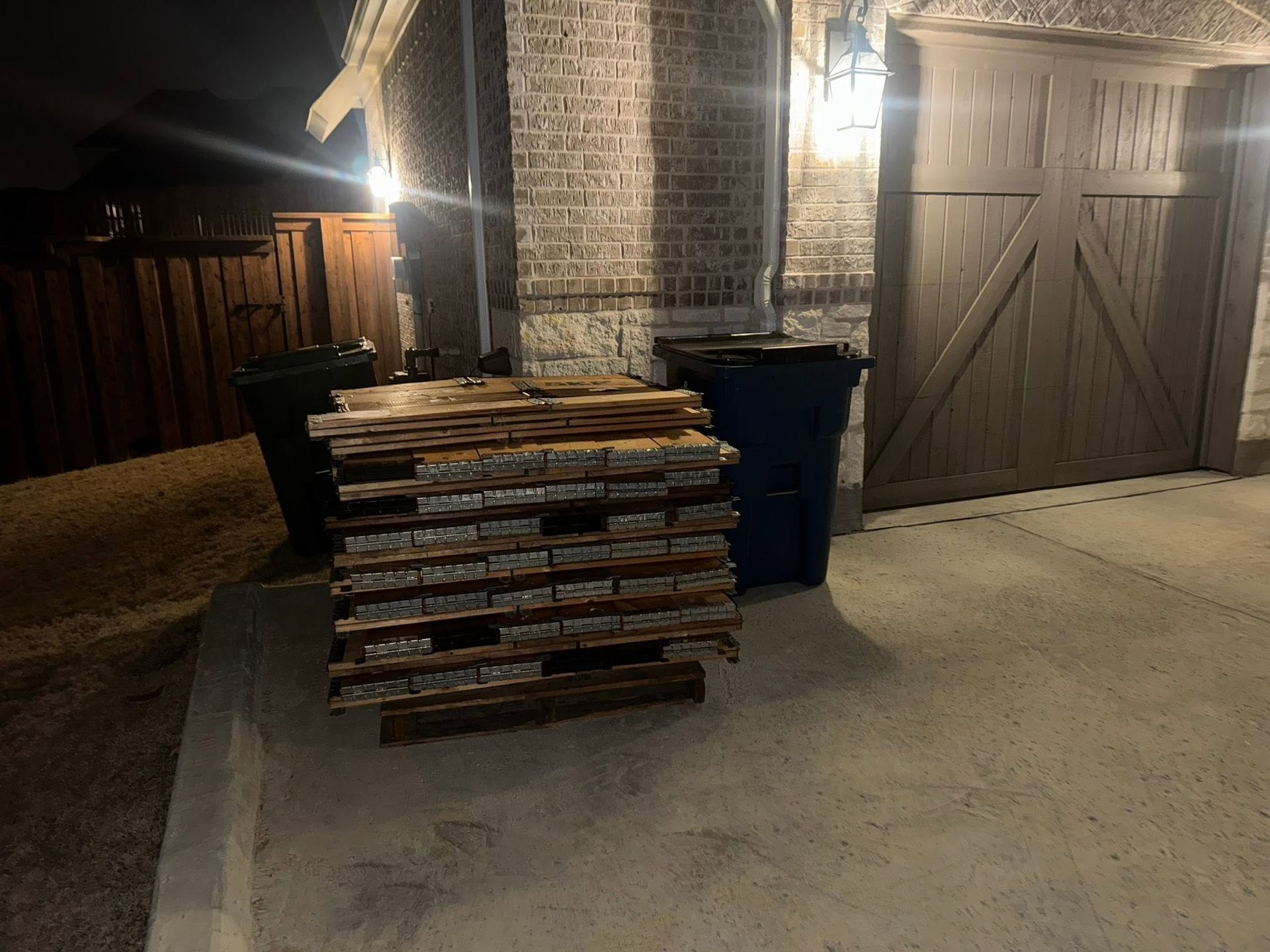 Stacked lumber in front of blue trash bins beside a brick house and garage at night