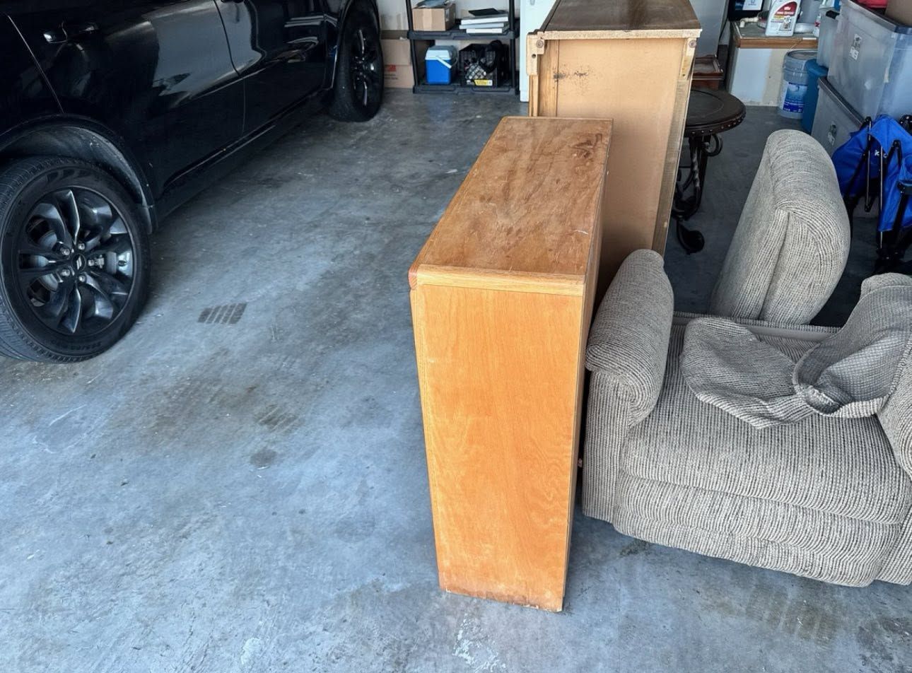 Wooden dresser standing in a cluttered garage beside a car and a gray upholstered chair.