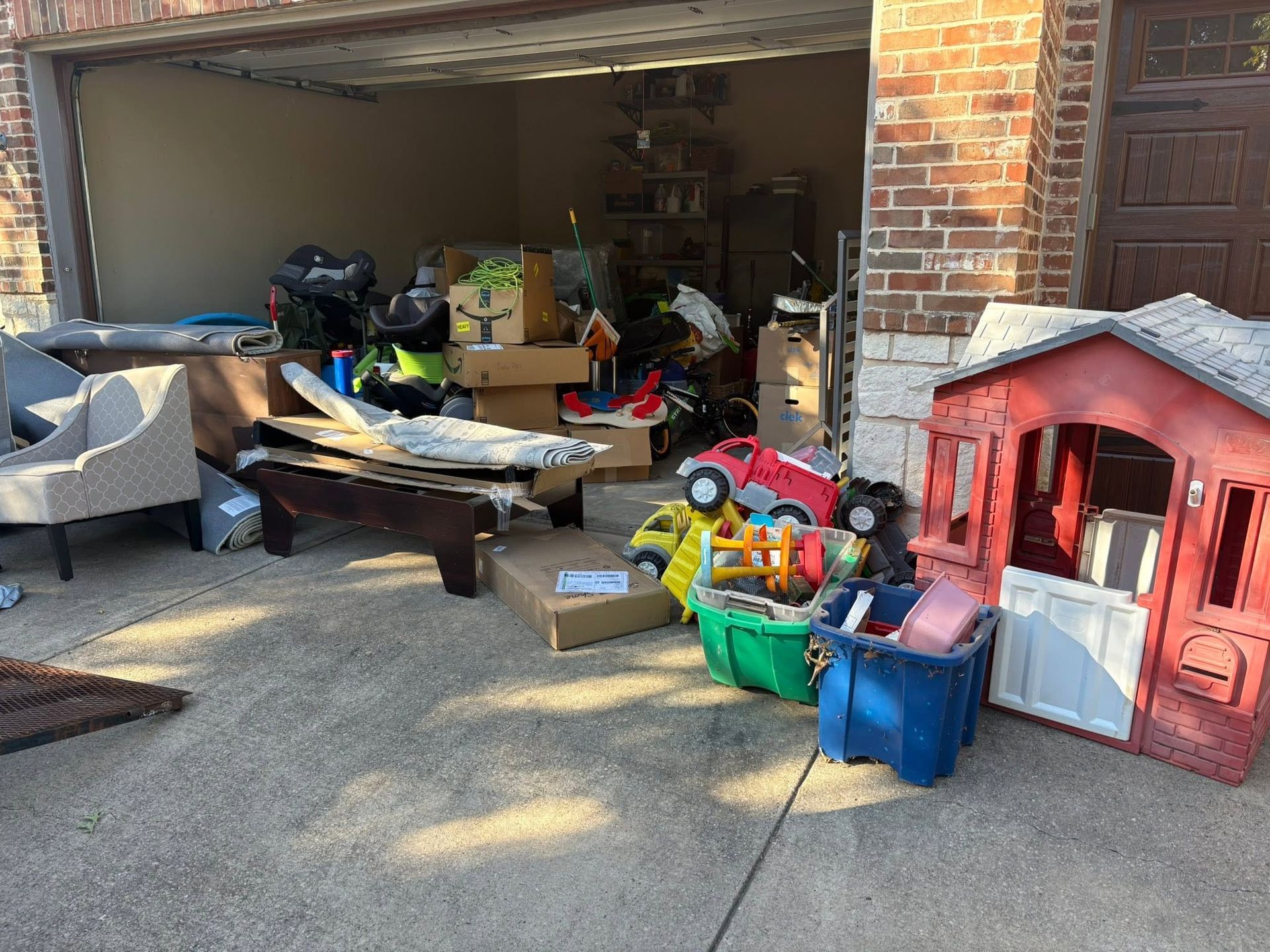 Cluttered garage with furniture, bags, toys, and a red child’s playhouse on the driveway
