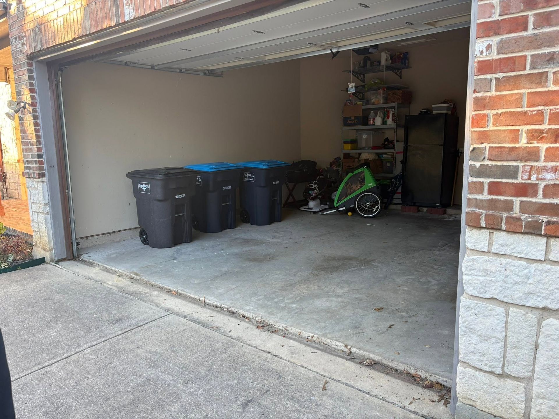Open garage with trash bins and a green child’s bike inside, concrete floor and brick exterior.