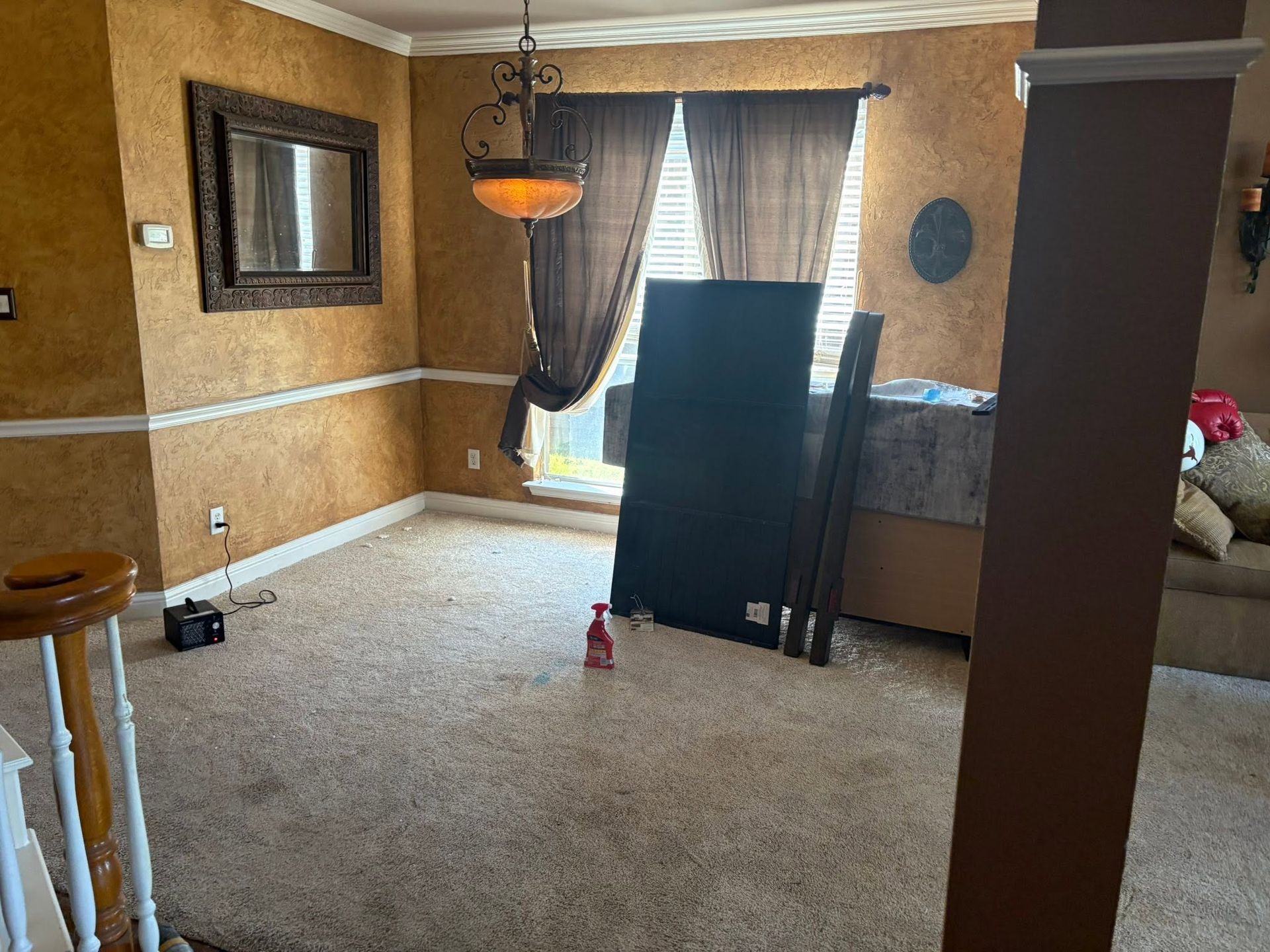 Empty living room with tan walls, carpet, window curtains, and a large TV near the center.
